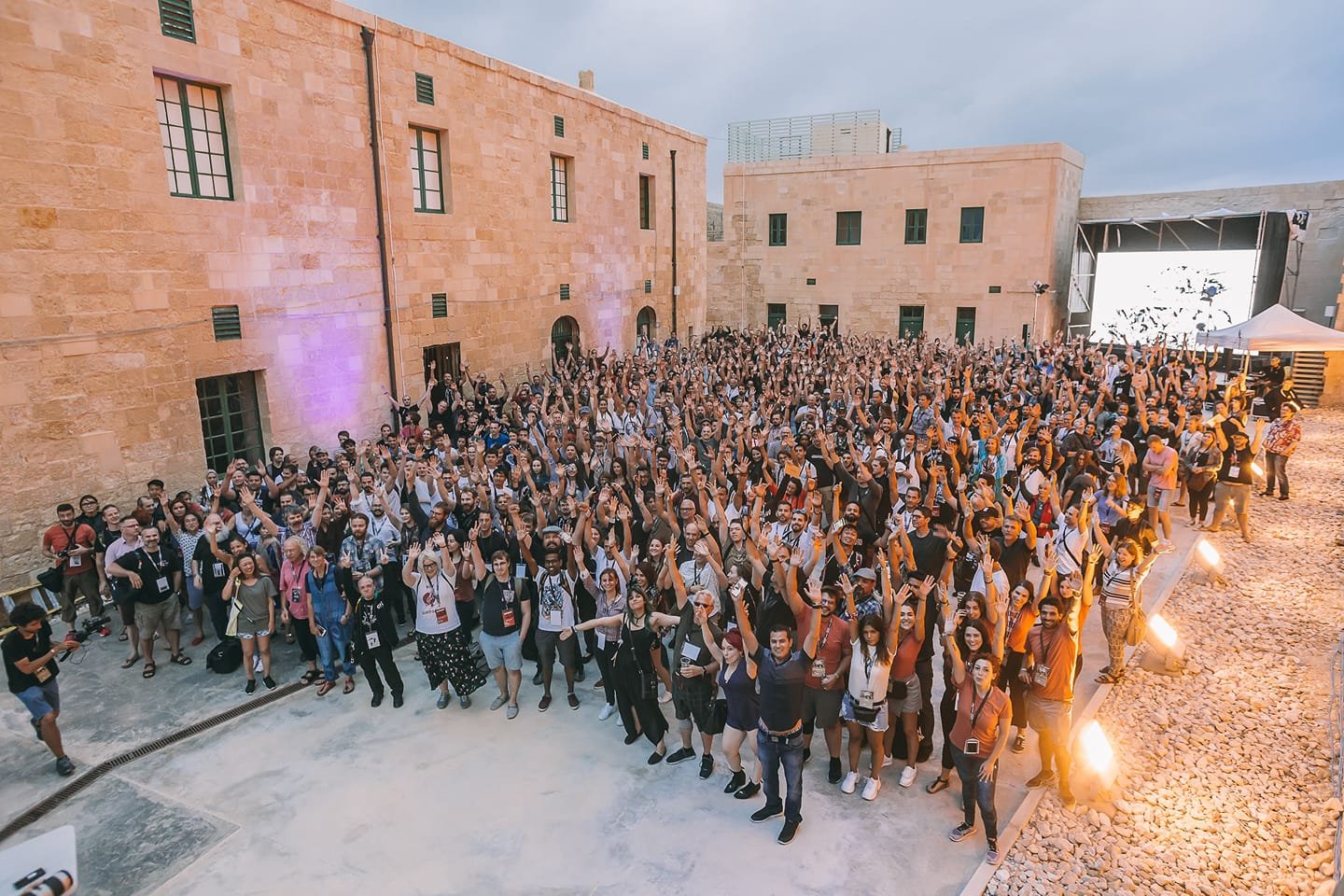 Large group of people gathered outdoors at an event, raising their hands and smiling, with a stage and large screen visible in the background.