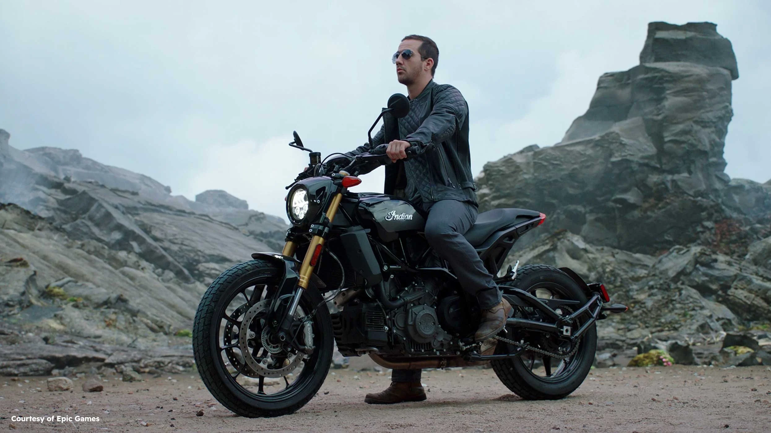 A man in sunglasses and a black leather jacket is sitting on a black Indian motorcycle on a rocky landscape with large rock formations in the background.