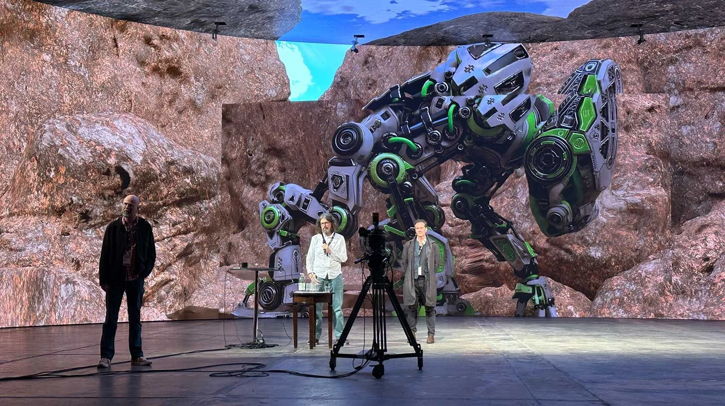 A large stage with a background image of a rocky landscape features a giant, realistic robotic creature resembling a dinosaur. Three men are on stage, one with a microphone, while a camera on a tripod captures the scene.