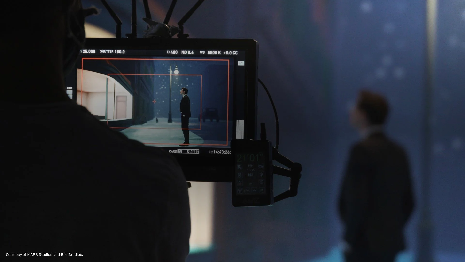 A film director on set monitors a scene of a man standing under a street lamp at night, seen through a camera viewfinder, with a blurred figure in the background.