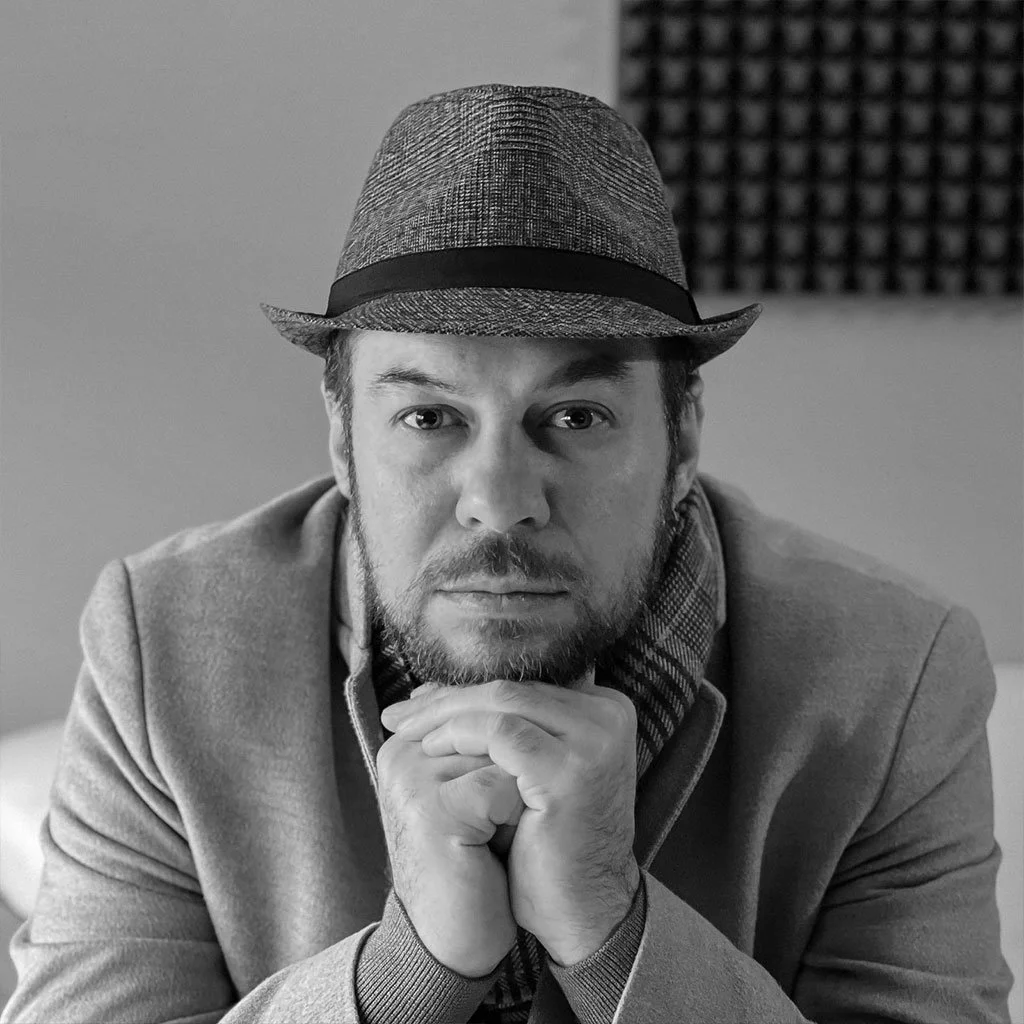 A man wearing a fedora hat with a black band, resting his chin on his hands, looking directly at the camera in a black and white photo.