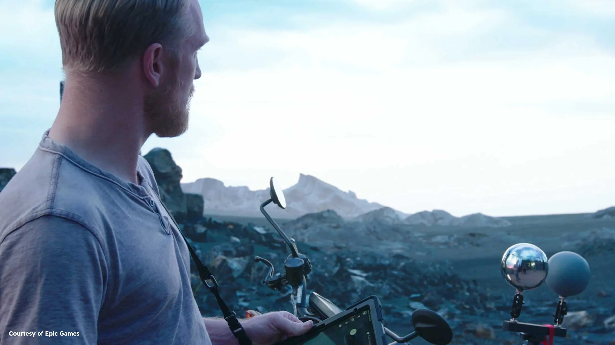 A man with red hair and a beard, wearing a light gray shirt, is operating a remote control with a tablet in an outdoor rocky landscape during daytime. There are two weather instruments mounted on a bike in front of him, one with a mirror-like reflective sphere and the other a matte sphere.