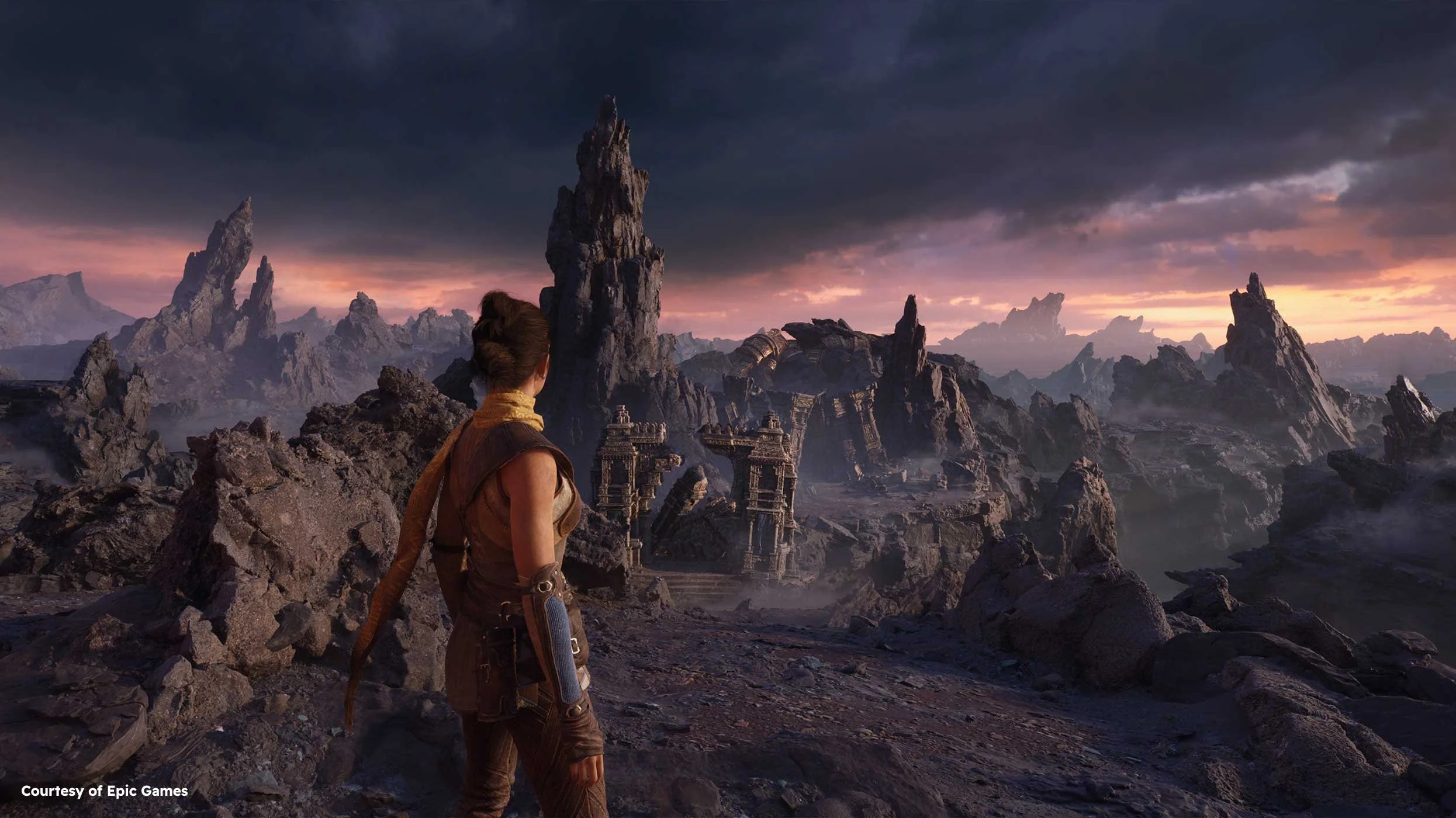 A woman with dark hair in a bun, wearing adventure gear, standing on rocky terrain and looking at ancient ruins in a mountainous landscape during sunset with dark clouds in the sky.