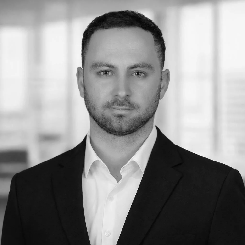 Black and white professional headshot of a man in a suit, with a serious expression, in an office setting.