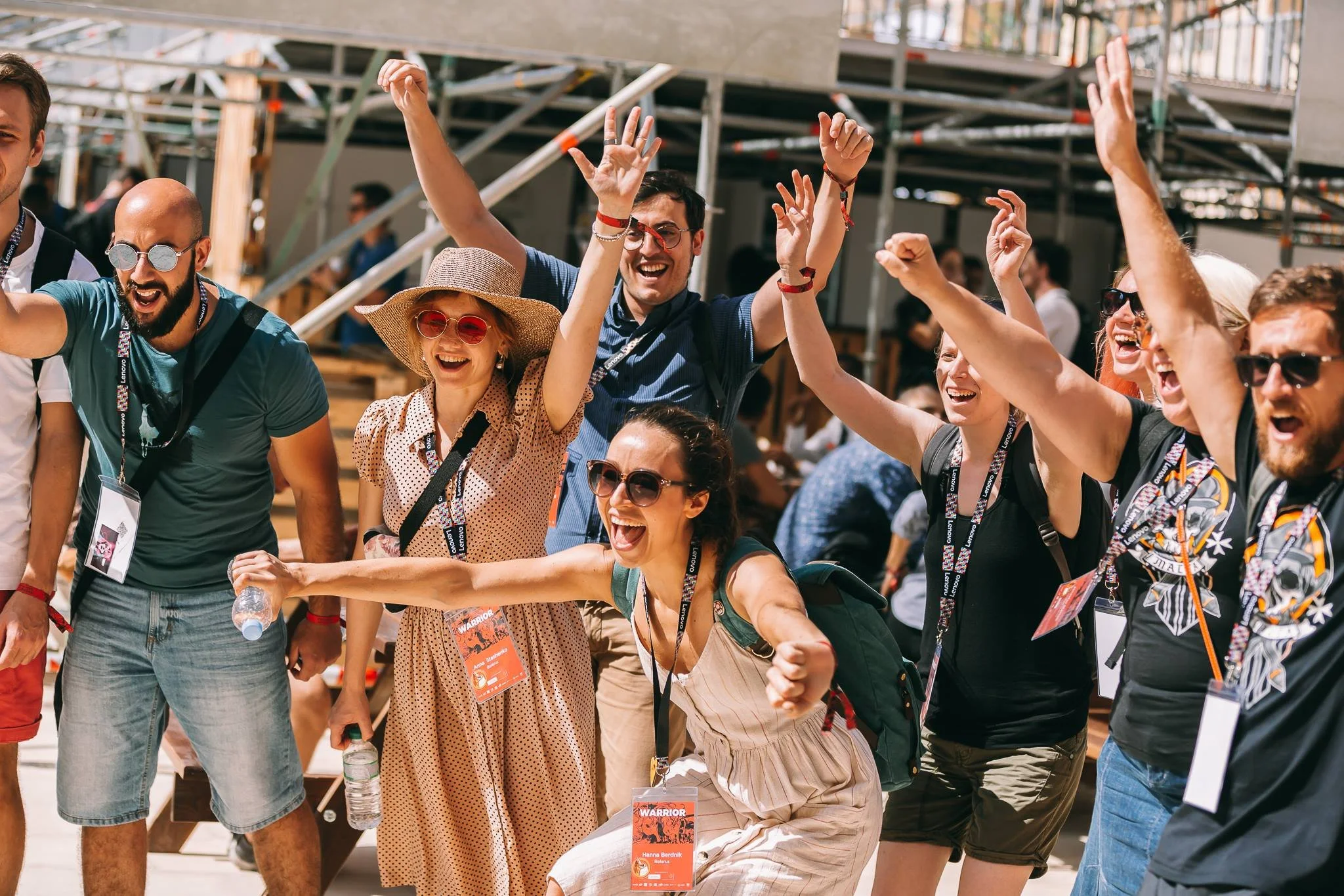 Group of happy, diverse young adults celebrating outdoors during the daytime at an event, wearing casual summer clothes, sunglasses, and festival badges, with some raising their hands and smiling.