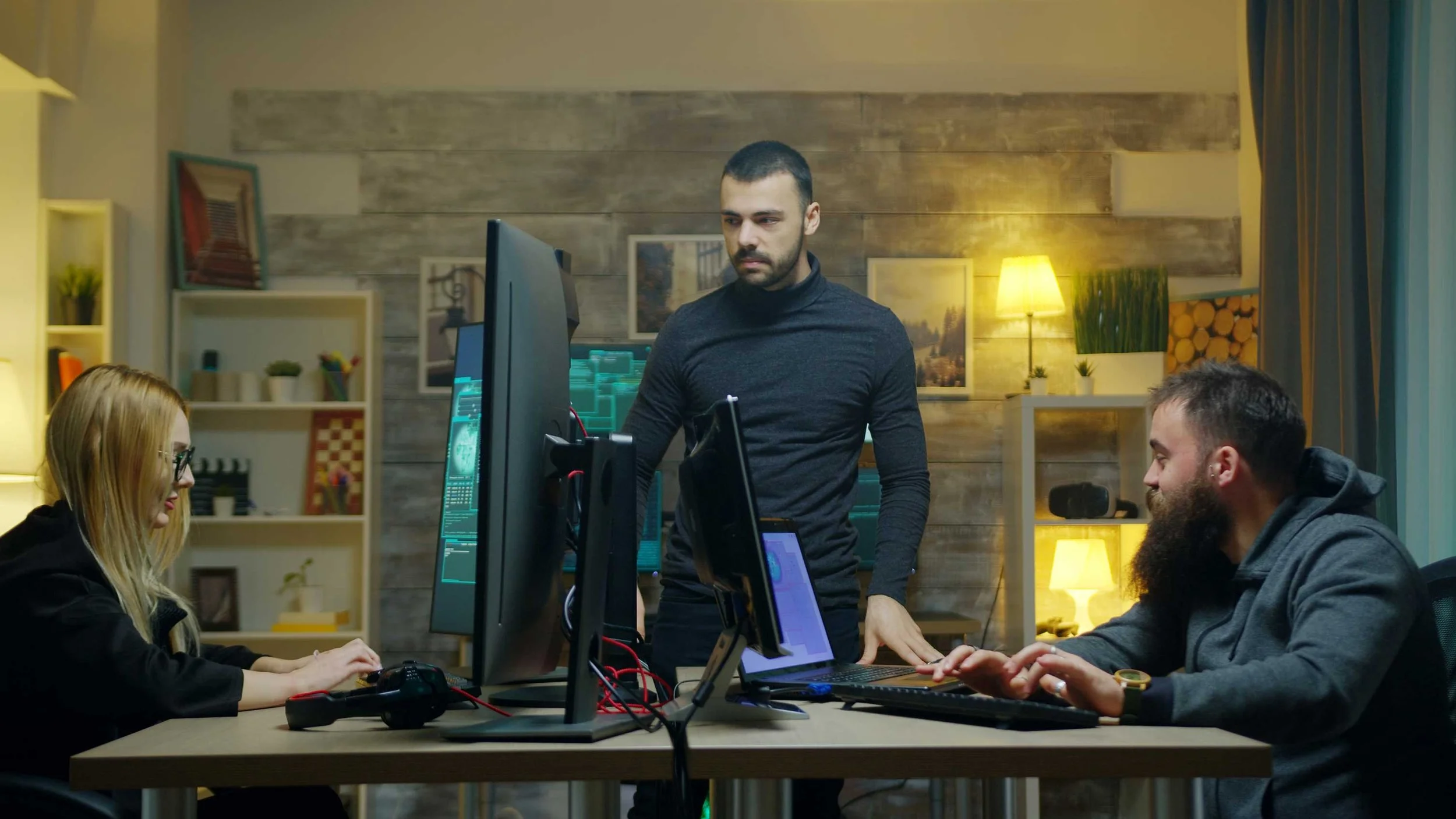 Three people working at a table with computer monitors in a cozy, well-lit room with bookshelves, pictures, and warm lighting.