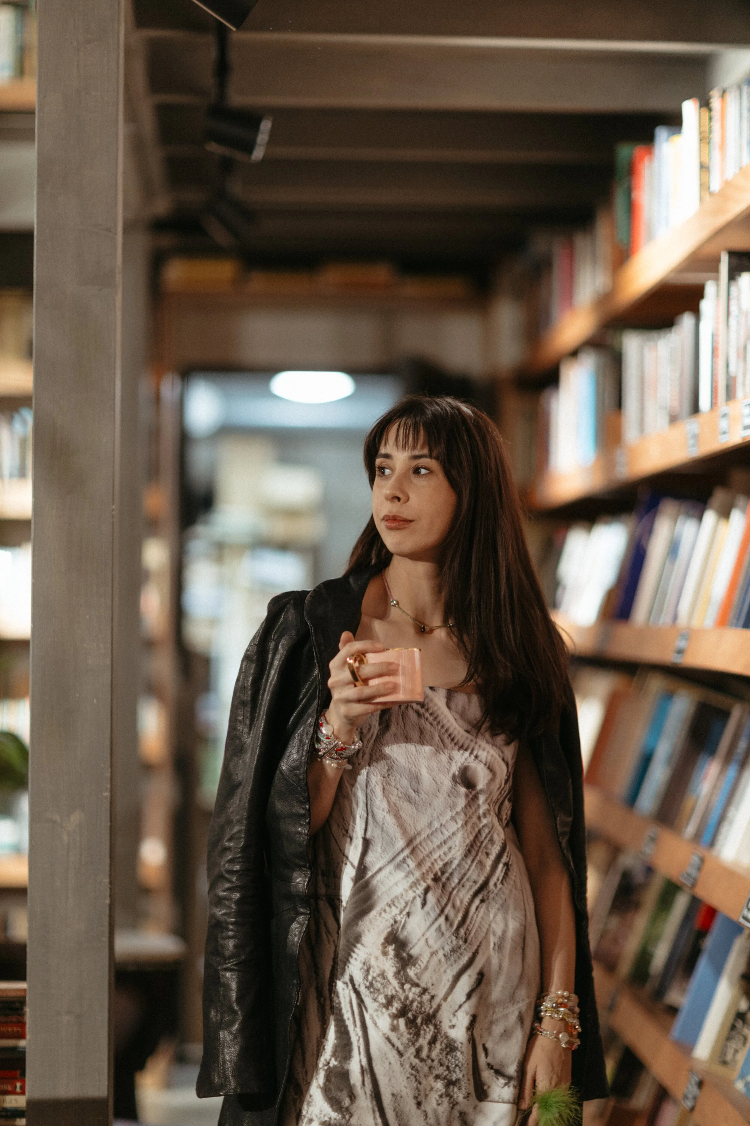 A woman with dark hair and bangs, wearing a printed dress and a black leather jacket, stands in a bookstore aisle holding a small cup, looking thoughtfully to the side.