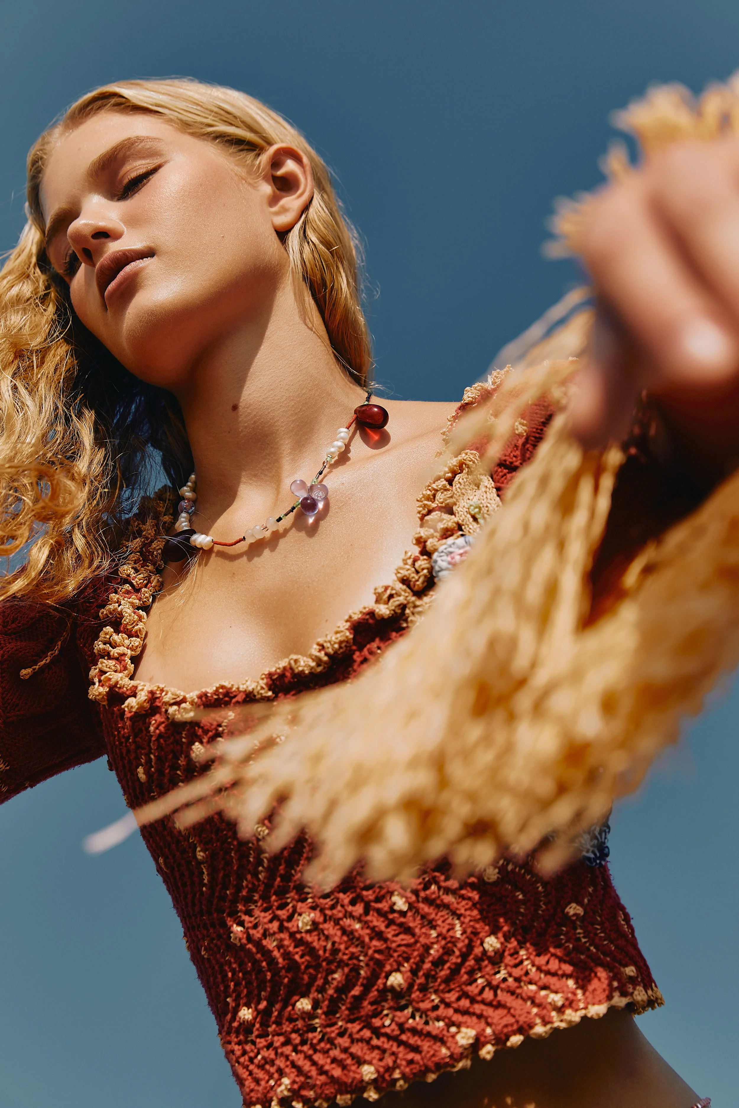 Close-up of a woman with blonde curly hair, wearing a red textured top with ruffled edges and a colorful beaded necklace, looking down with her eyes closed against a clear blue sky.