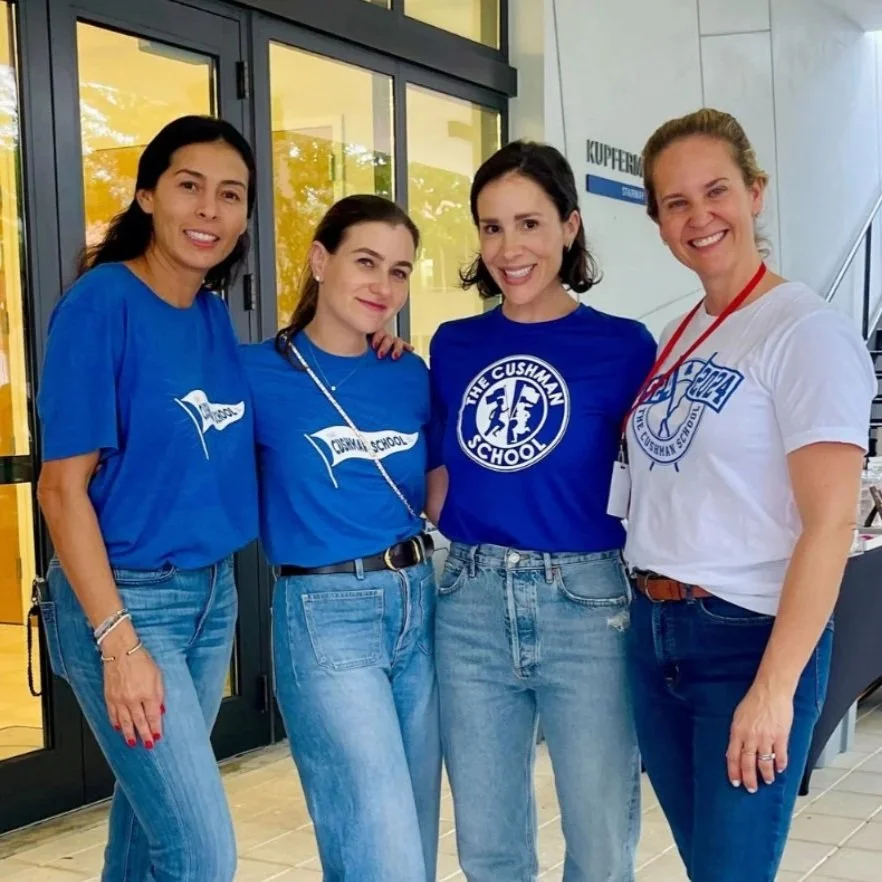Four women standing side by side in front of a glass door, all smiling and wearing shirts with school logos, celebrating or attending a school event.