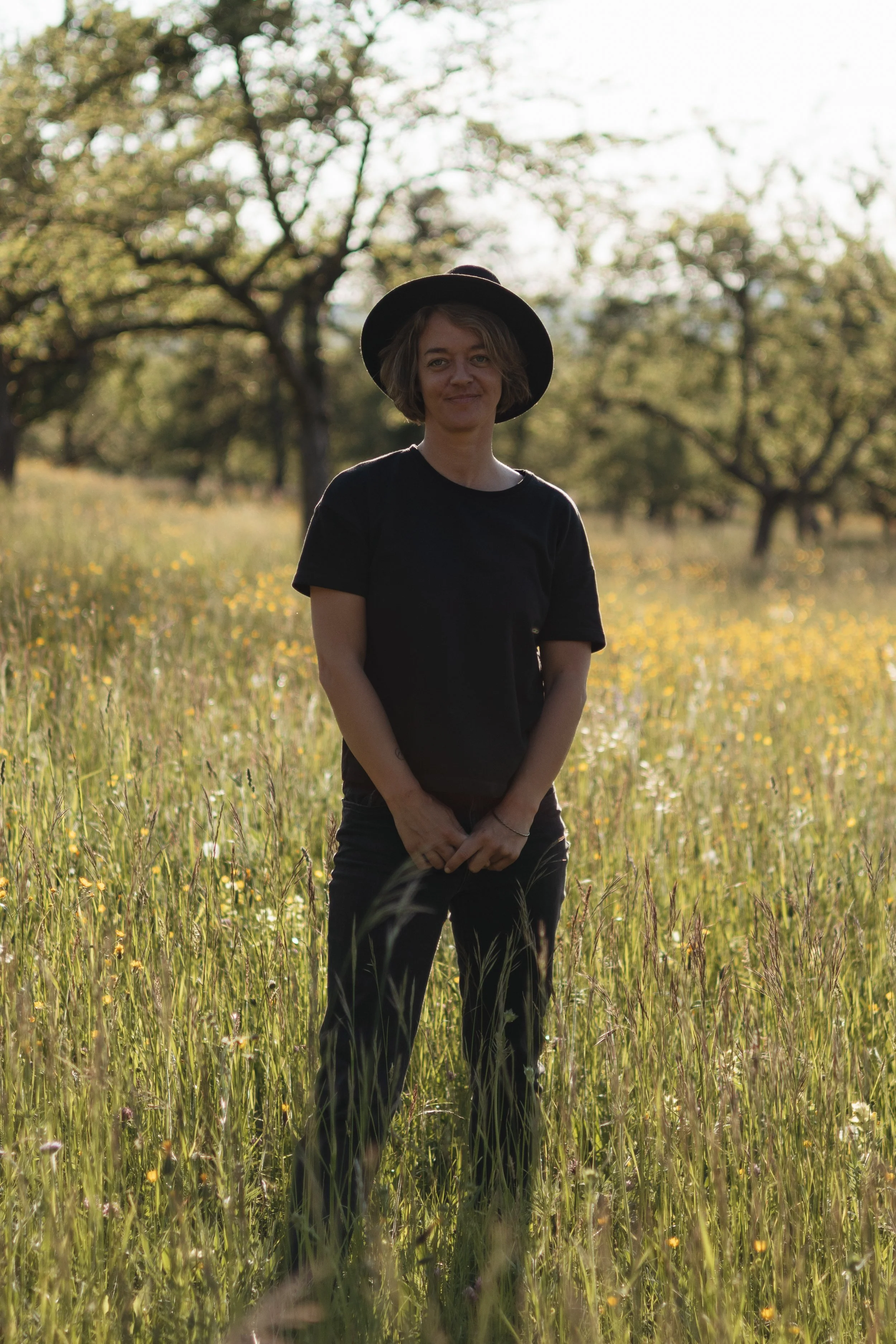 Eine Frau trägt einen schwarzen Hut und schwarzen T-Shirt im Sonnenlicht in einem Feld mit gelben Blumen, im Hintergrund Bäume.