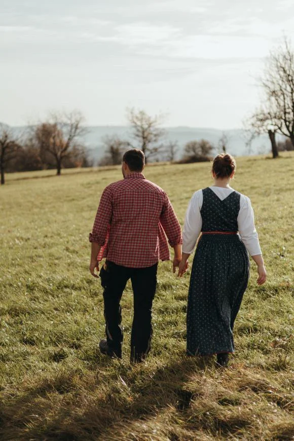 Ein Paar in traditioneller Kleidung spaziert in einem offenen, grünen Feld mit Bäumen im Hintergrund bei Sonnenuntergang.