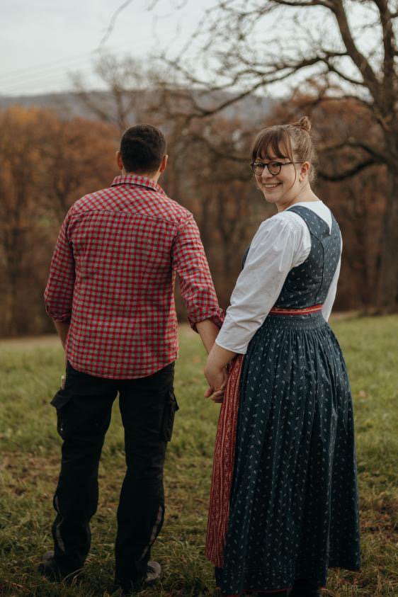 Ein Paar steht im Freien, hält sich an den Händen und blickt sich gegenseitig an. Die Frau trägt ein traditionelles Kleid und die Herbstlandschaft im Hintergrund.