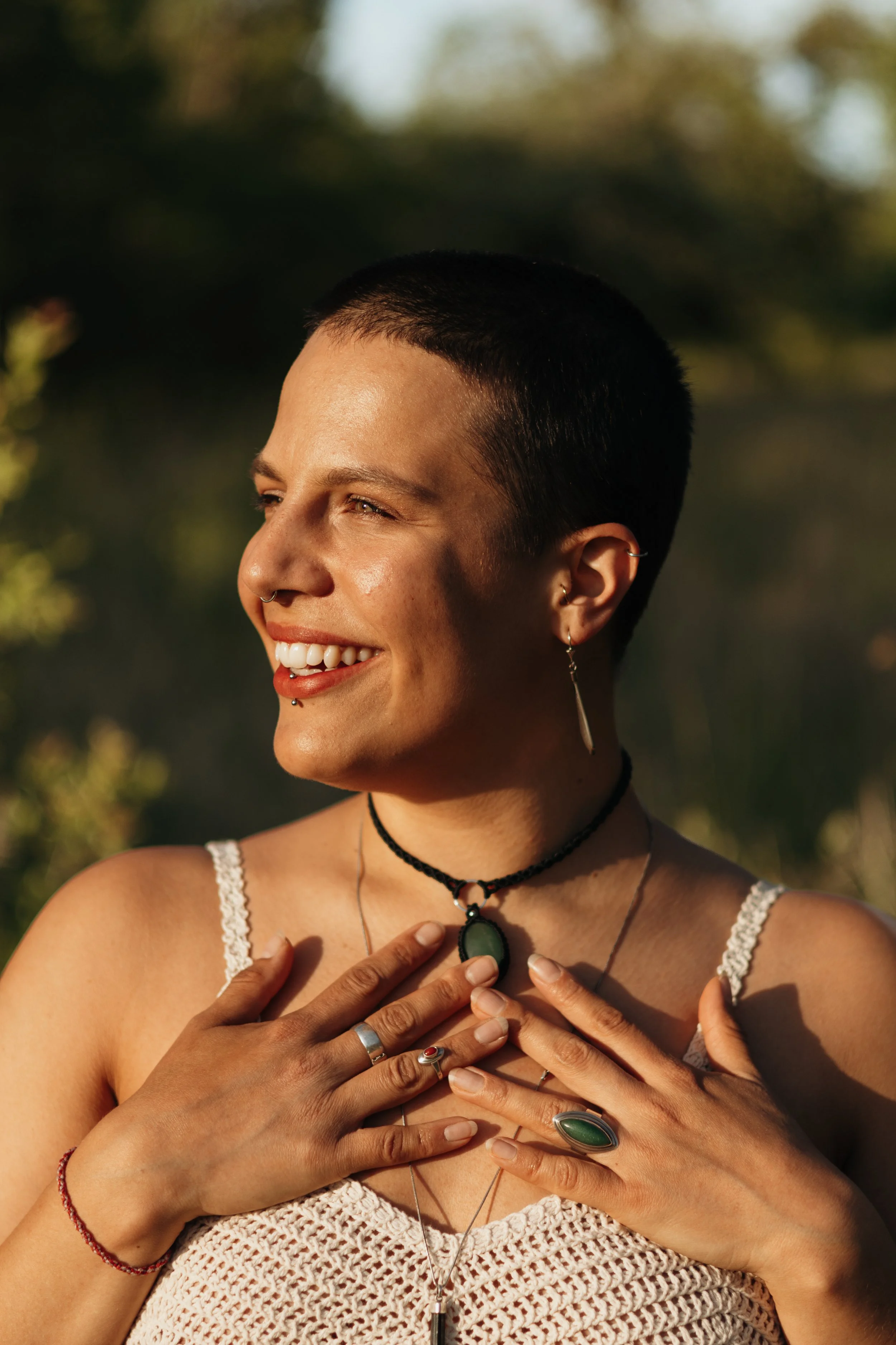 Eine lachende Frau mit kurzen dunklen Haaren, mehreren Piercings, Schmuck und einer weißen, häkelartigen Top im Freien bei Sonnenuntergang.