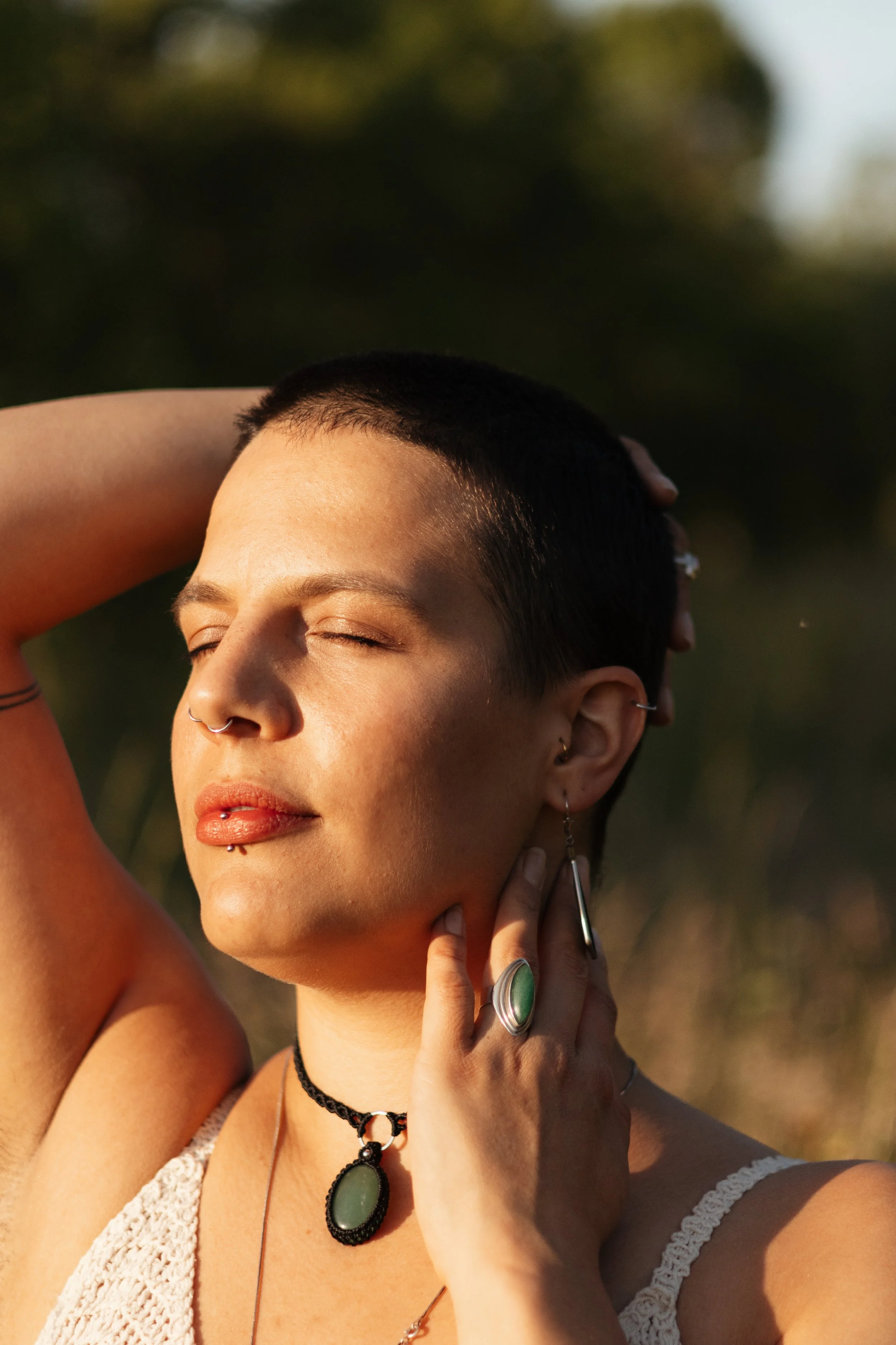 Eine Person mit kurzen dunklen Haaren und mehreren Piercings trägt Schmuck aus grünen Steinen. Sie steht mit geschlossenen Augen im Sonnenlicht, umgeben von einem natürlichen Hintergrund.