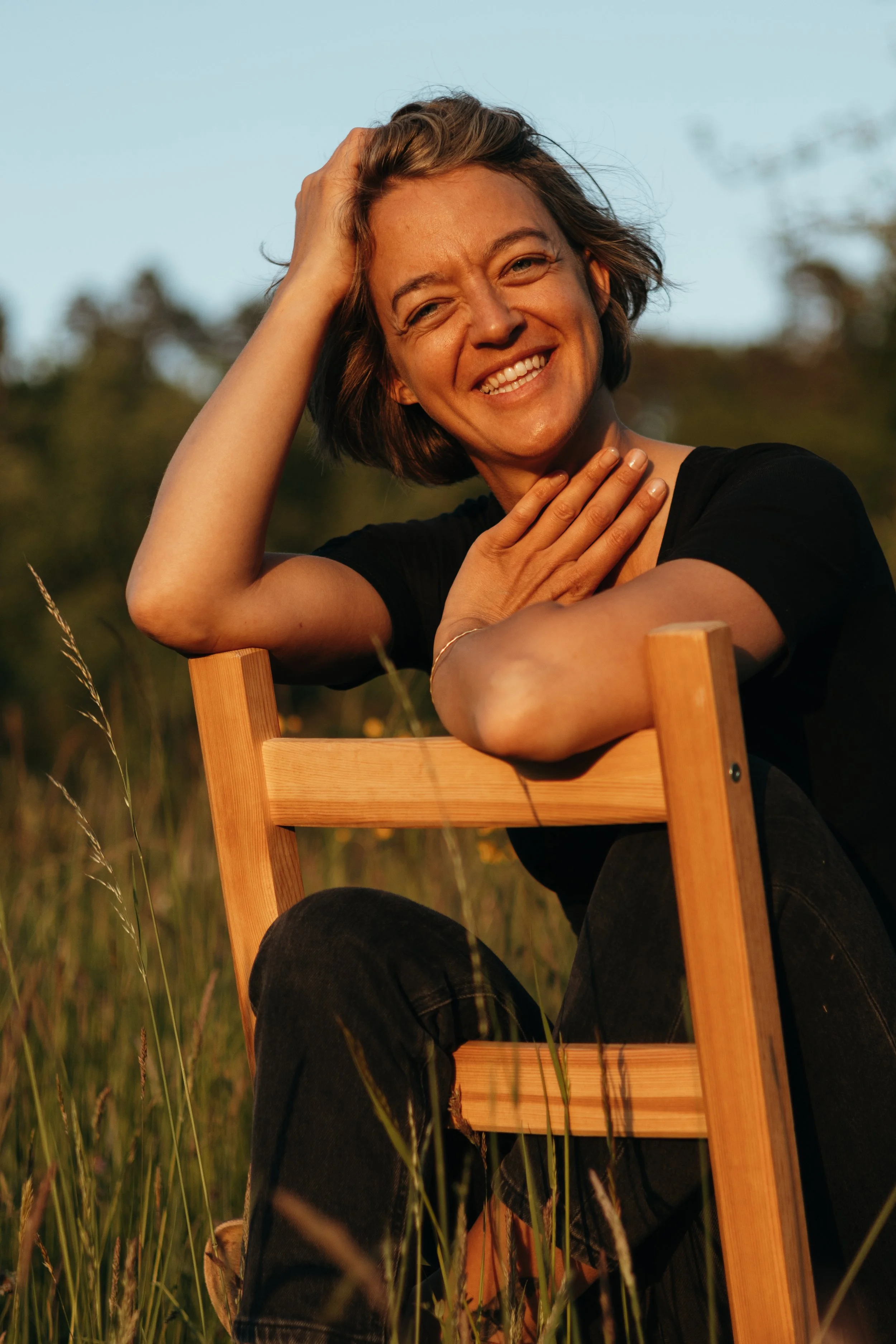 Lächelnde Frau sitzt im Freien auf einem Holzstuhl in einem Feld bei Sonnenuntergang.