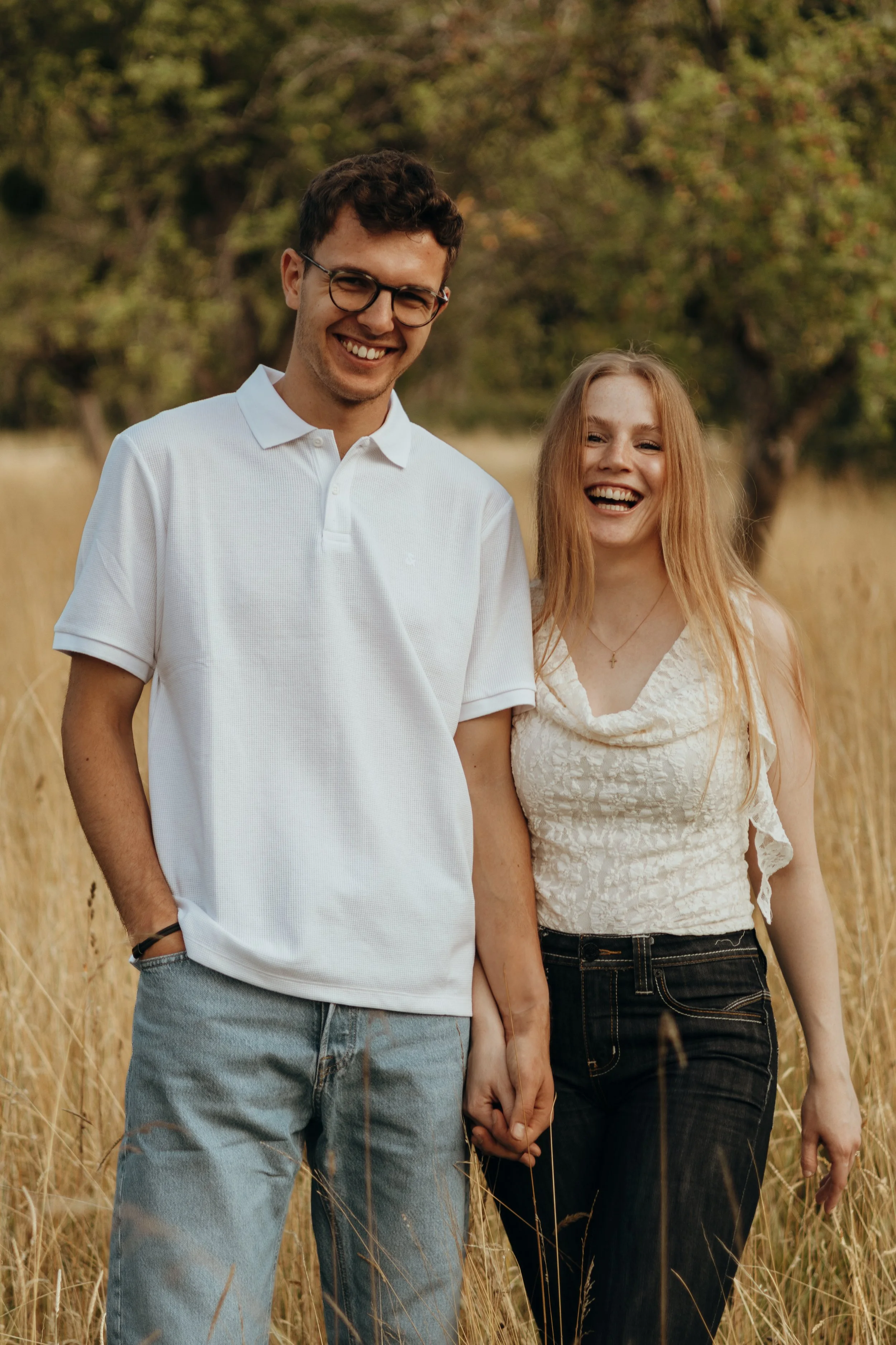 Ein junges Paar steht Hand in Hand in einem Feld mit hohen Gräsern, umgeben von Bäumen, lächelnd im Sonnenschein.