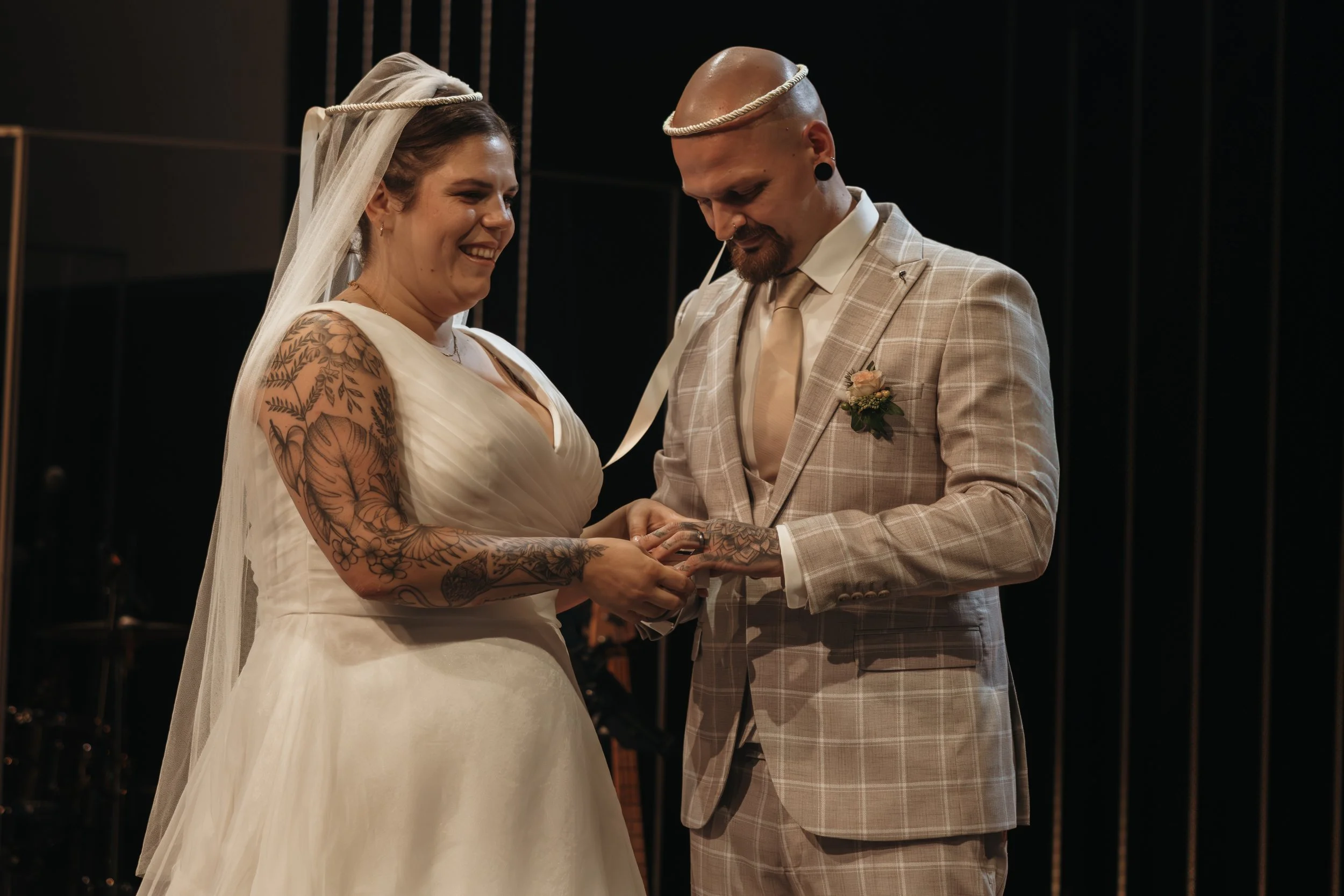 Ein Brautpaar bei einer Zeremonie, der Bräutigam steckt dem Braut eine Ringe an, beide lächeln.