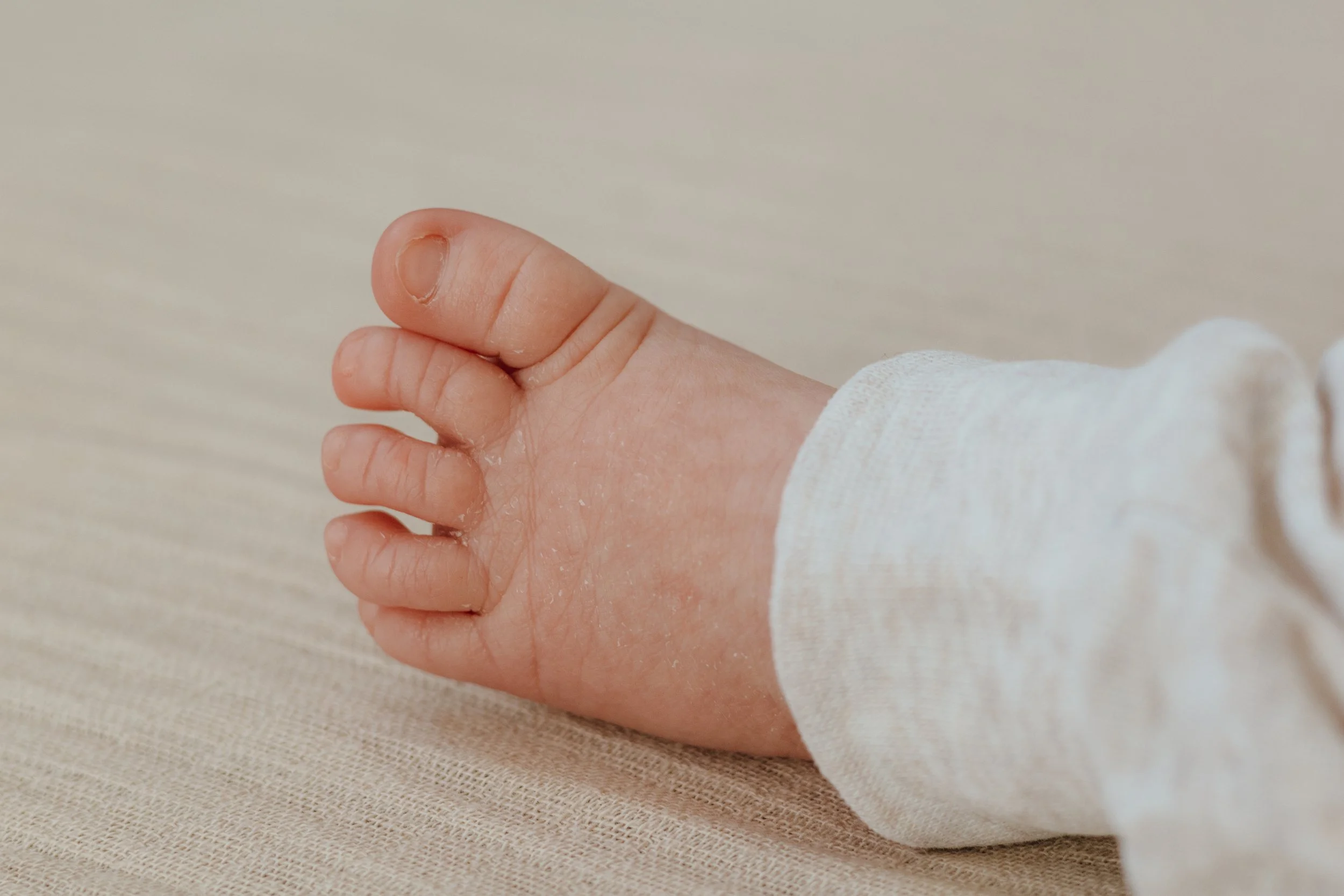Nahaufnahme eines kleinen Kinderfingers, der auf einer beige Stoffoberfläche liegt, teilweise bedeckt von einem weißen Langarmshirt.