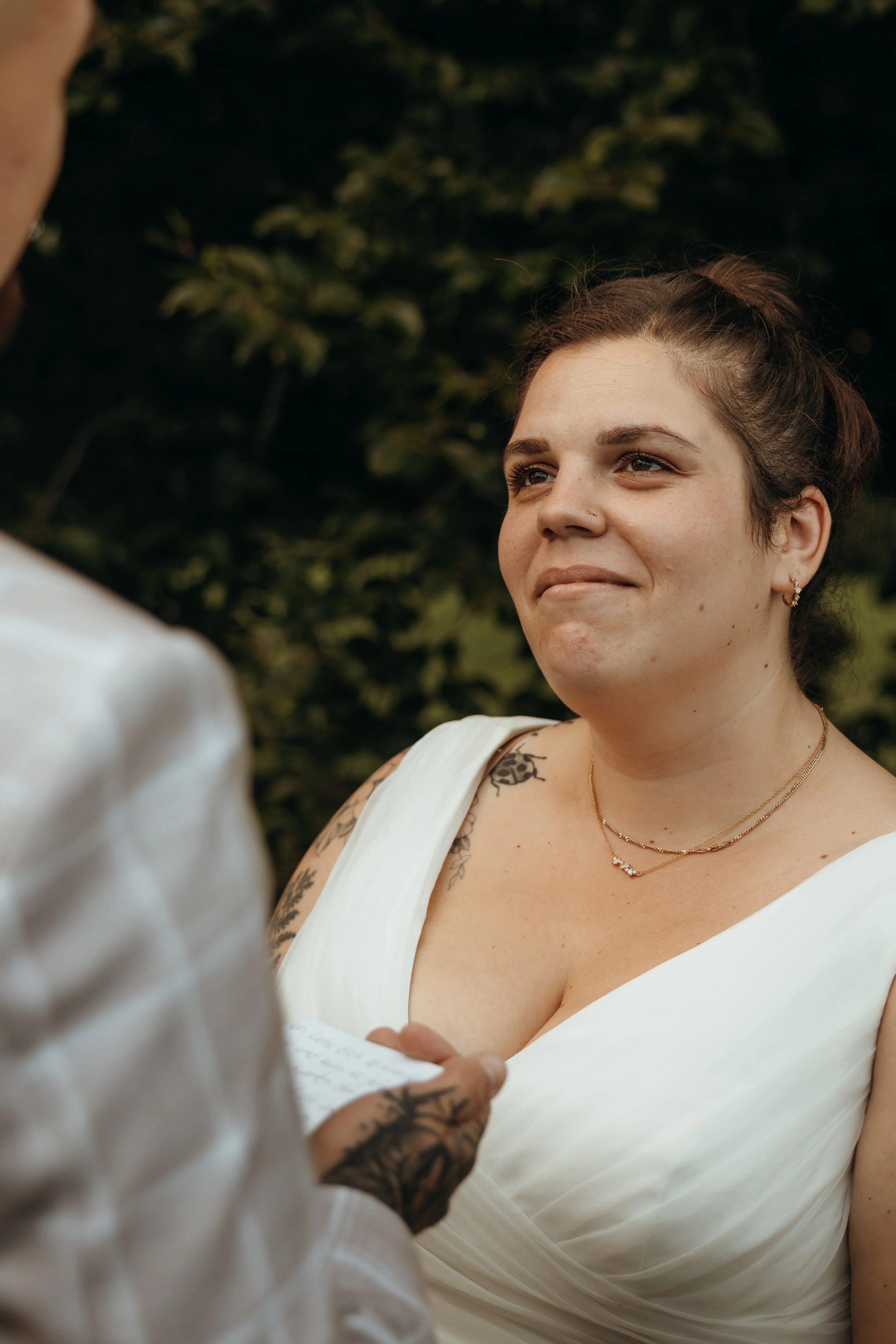 Eine junge Frau bei einer Zeremonie, vermutlich bei ihrer Hochzeit, trägt ein weißes Kleid, hat Tattoos am Arm und eine Halskette. Sie schaut emotional zu jemandem, der eine Notiz liest.