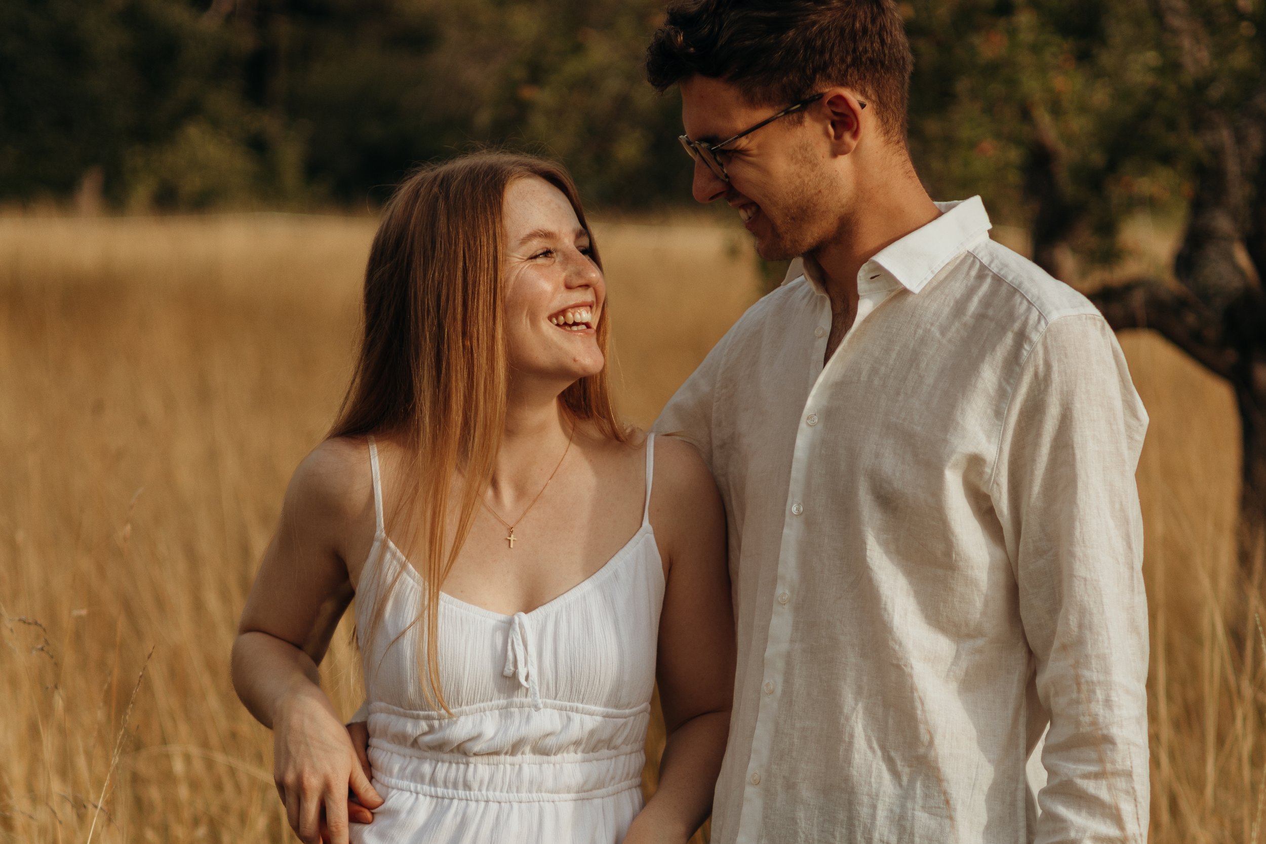 Ein lachendes Paar, ein Mann und eine Frau, stehen sich in einem Feld mit hohen Gräsern gegenüber, umgeben von Bäumen, bei Sonnenuntergang.