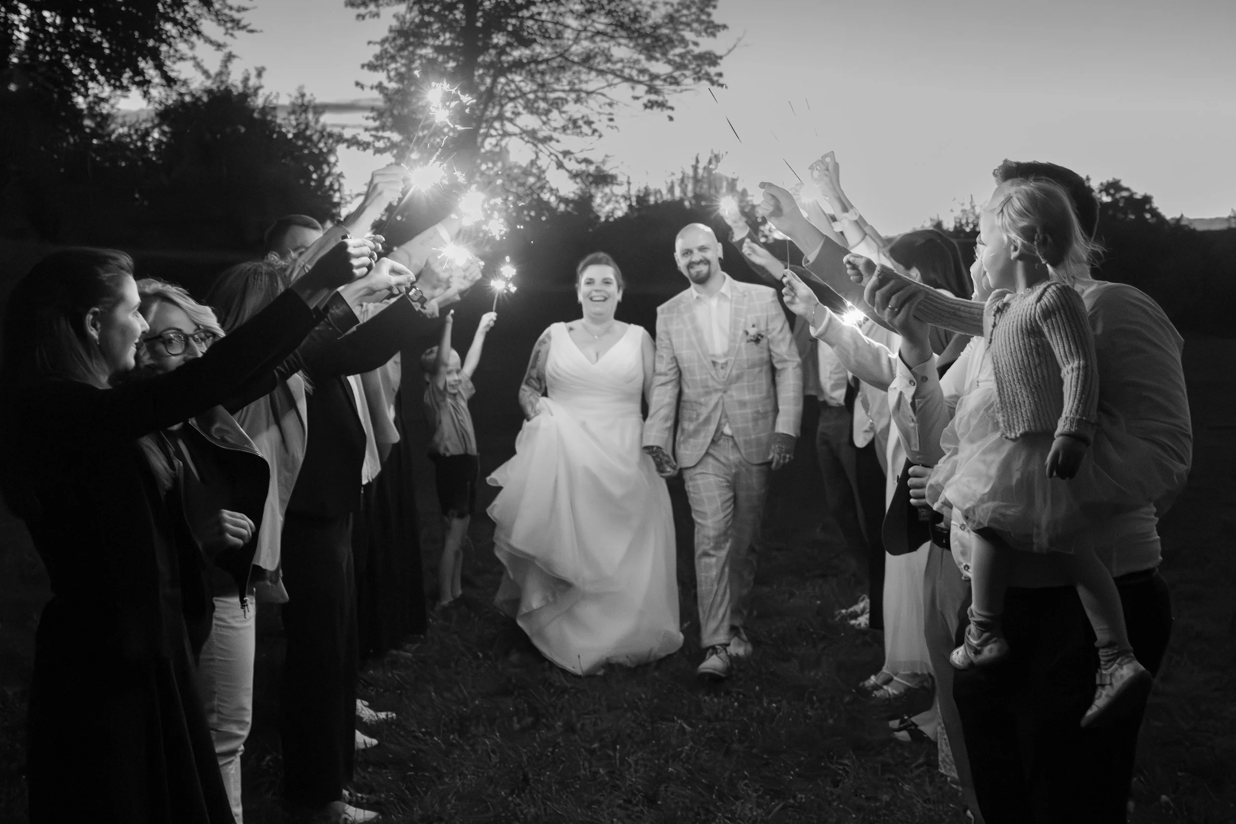 Ein Brautpaar läuft lachend und glücklich durch eine Gasse aus Freunden und Familienmitgliedern, die mit Wunderkerzen feiern, bei Sonnenuntergang im Freien.