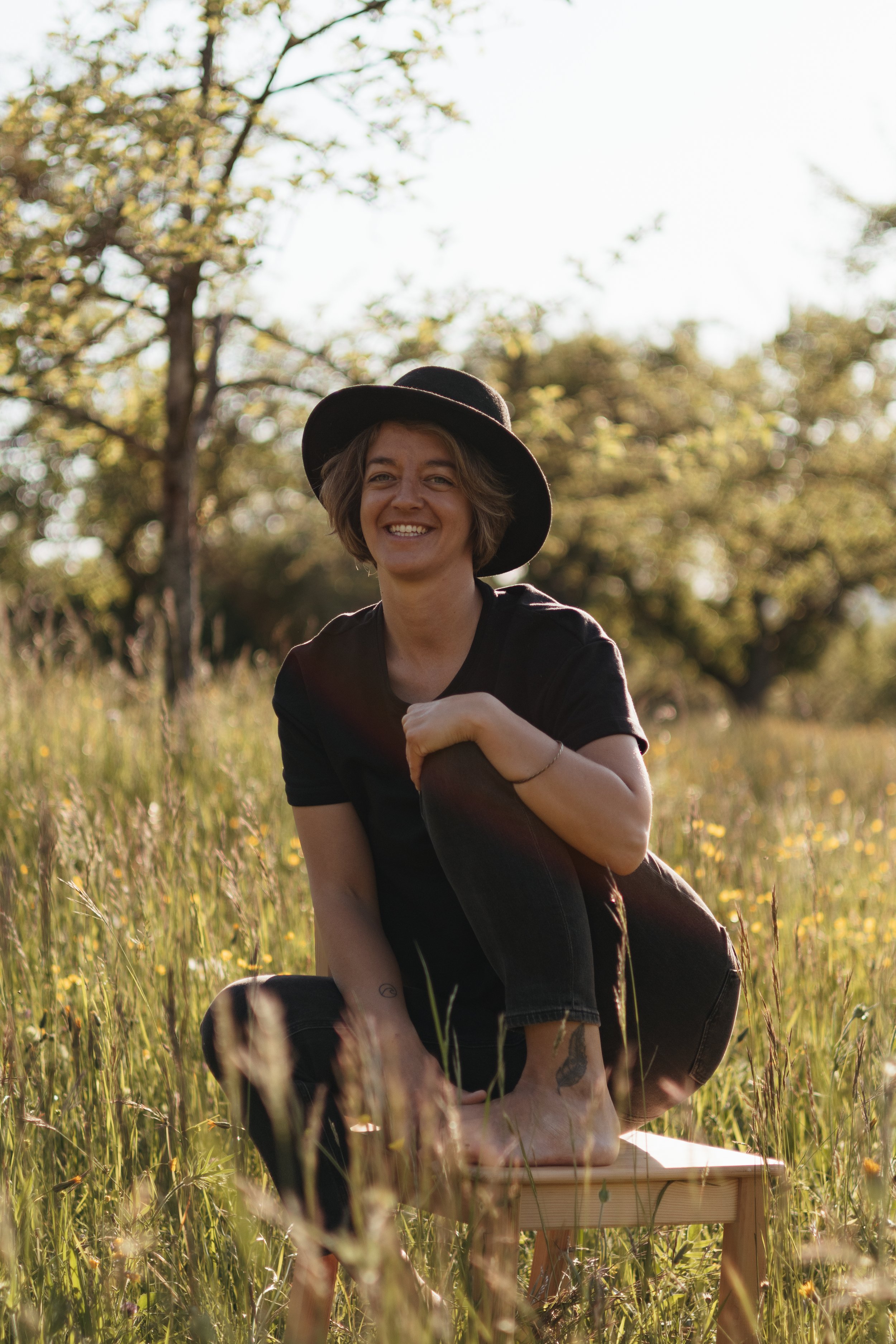Junge Frau mit Hut sitzt im Gras auf einer Wiese mit Bäumen im Hintergrund, bei Sonnenuntergang, lachend.