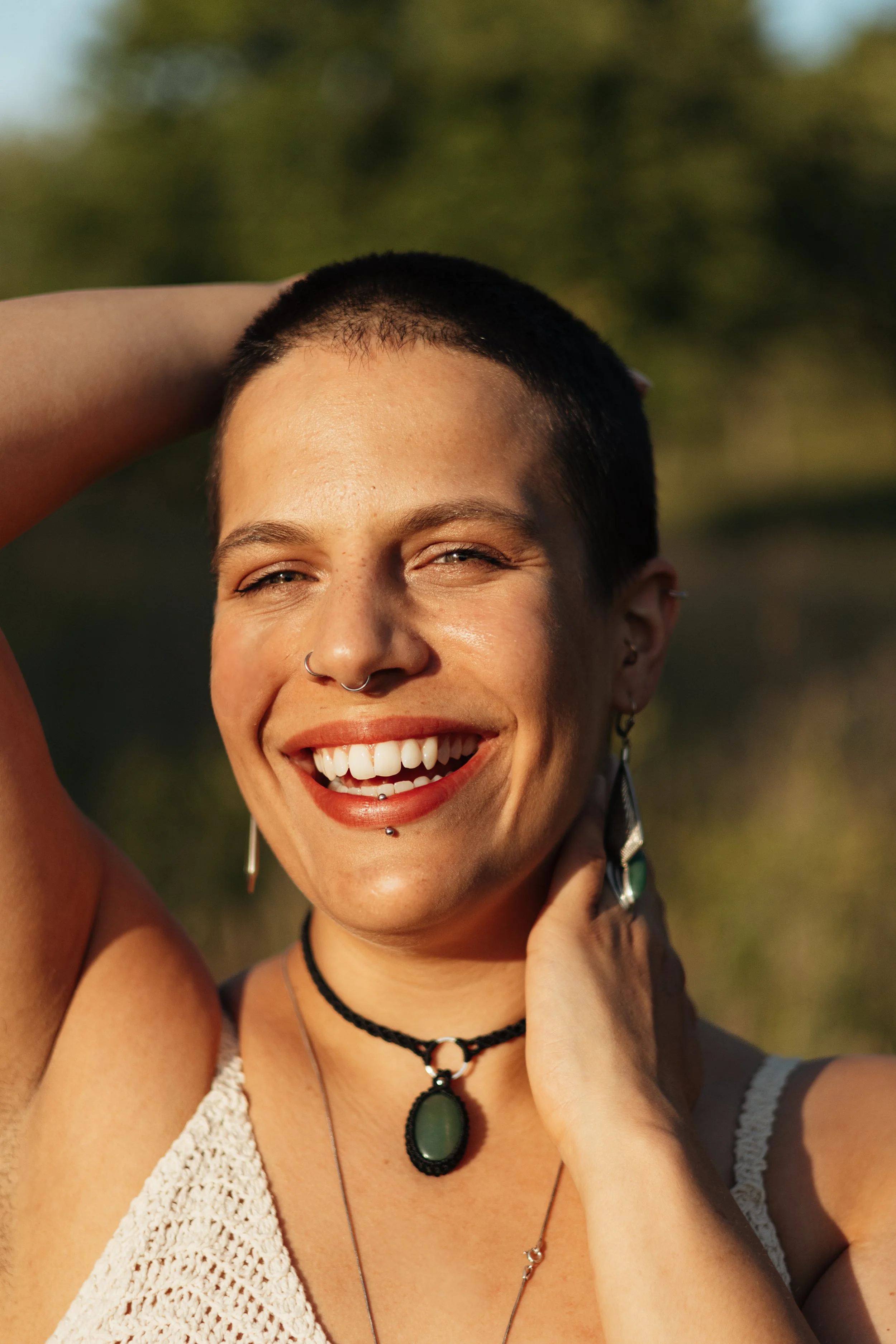 Junge Frau mit Kurzhaarschnitt, Piercings, lacht in die Kamera, im Freien bei Sonnenuntergang, trägt Schmuck, Sommerkleidung.
