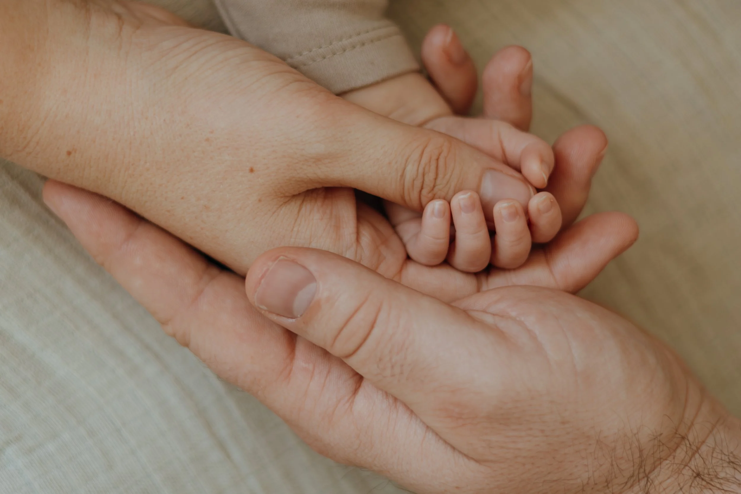 Eine erwachsene Hand hält eine kleine Kinderhand. Beide Hände liegen auf einer beigefarbenen Oberfläche.