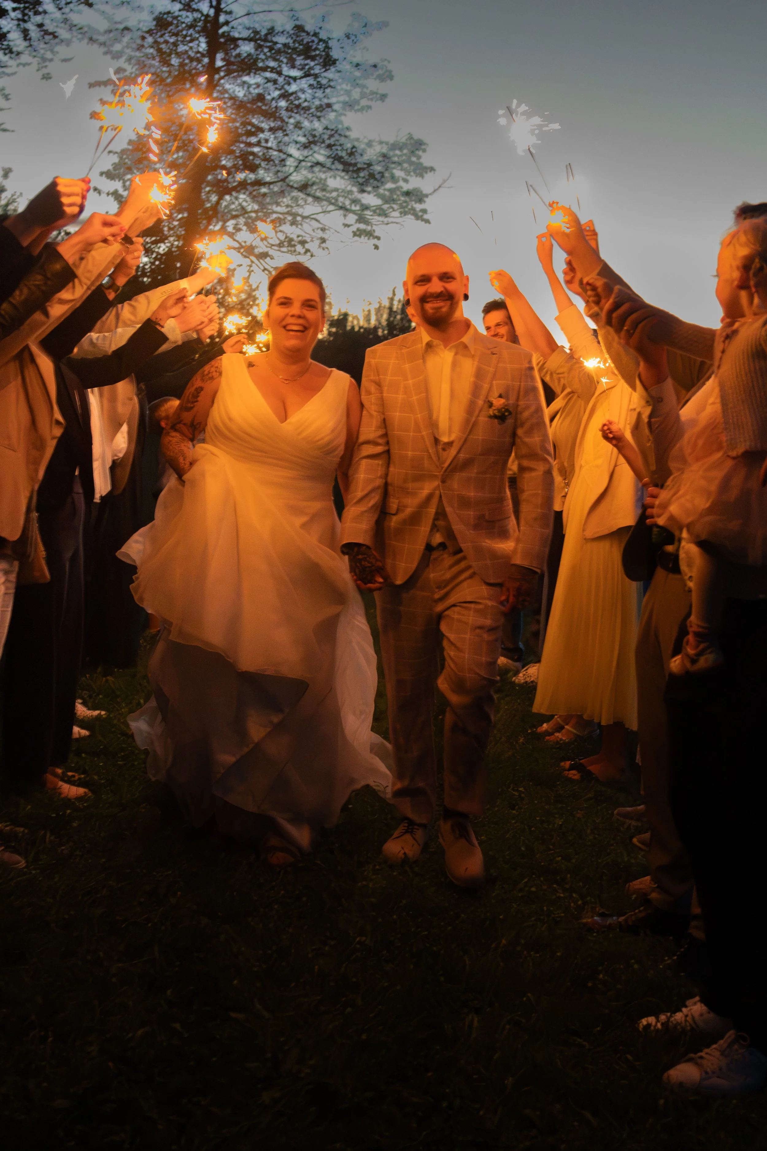 Ein glückliches Brautpaar läuft bei Sonnenuntergang durch einen Ring aus Gästen, die mit Kerzenhülsen leuchten, bei einer Hochzeitsfeier im Freien.