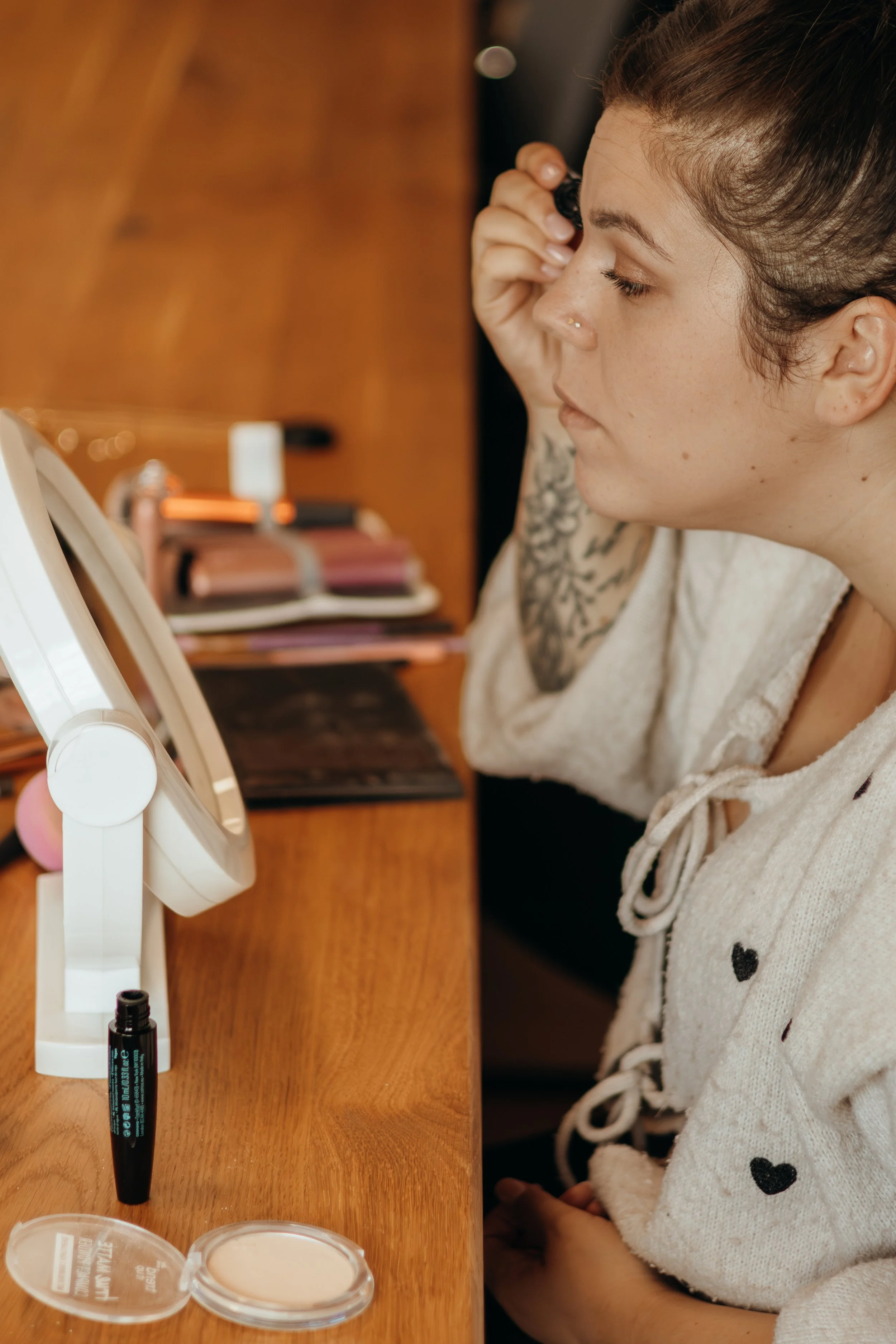 Junge Frau schaut in einen Spiegel beim Schminken, Makeup-Produkte und Pinsel auf dem Tisch.