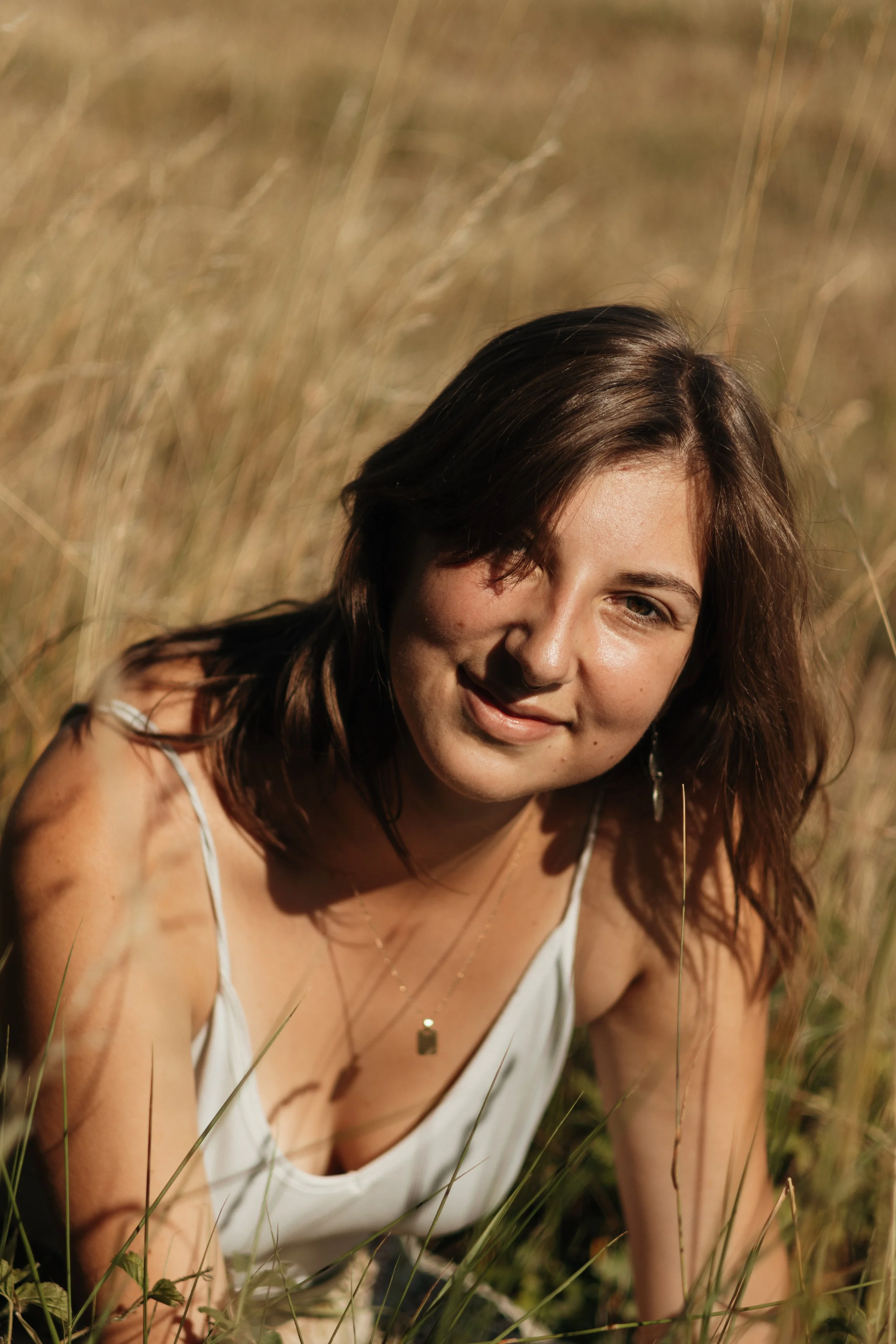 Junge Frau in einem weißen Kleid, die in einem Feld mit hohem Gras sitzt und in die Kamera lächelt.