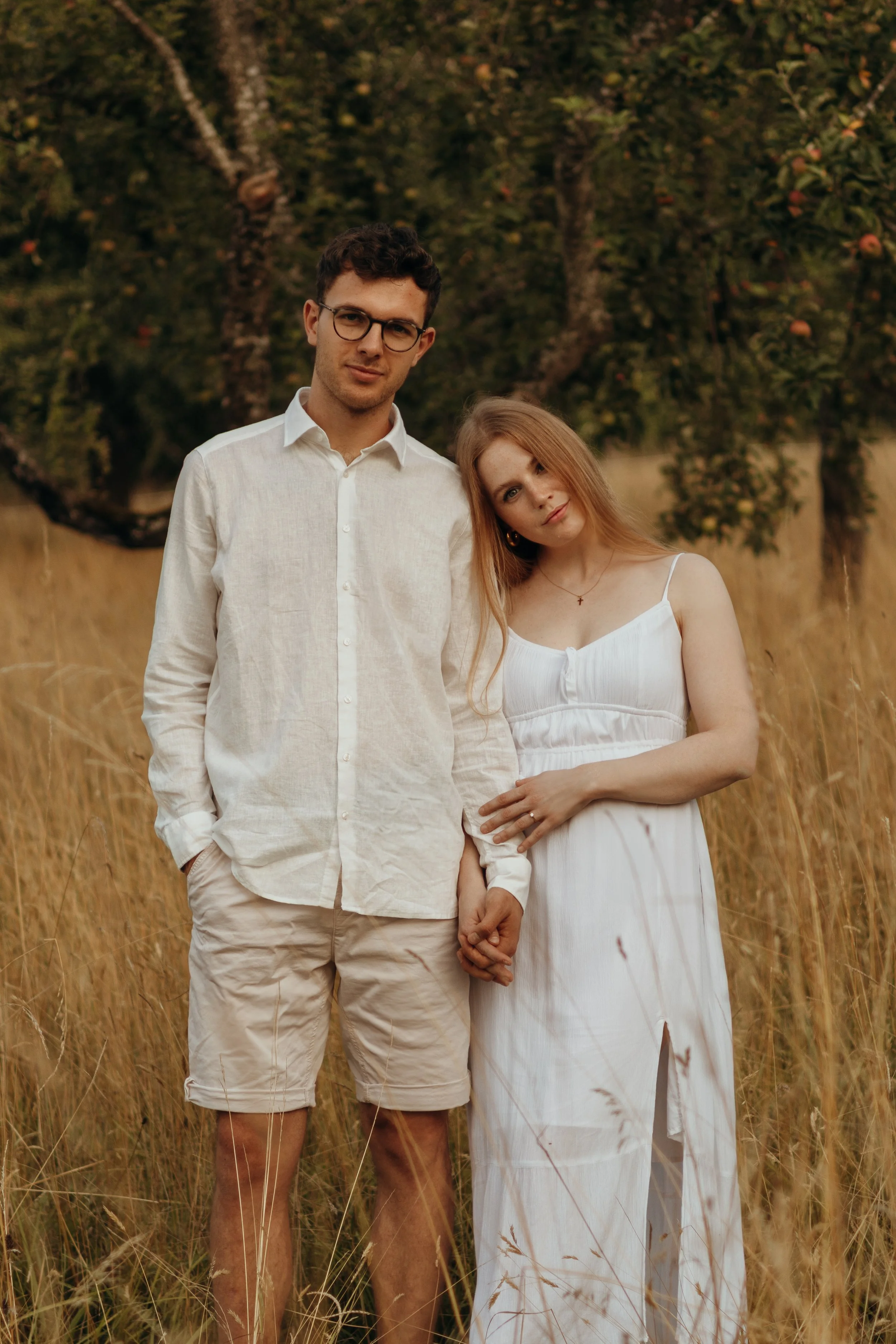 Ein junges Paar steht im hohen Gras vor einem Apfelbaum, Hand in Hand, die Frau trägt ein weißes Kleid, der Mann trägt helle Kleidung.