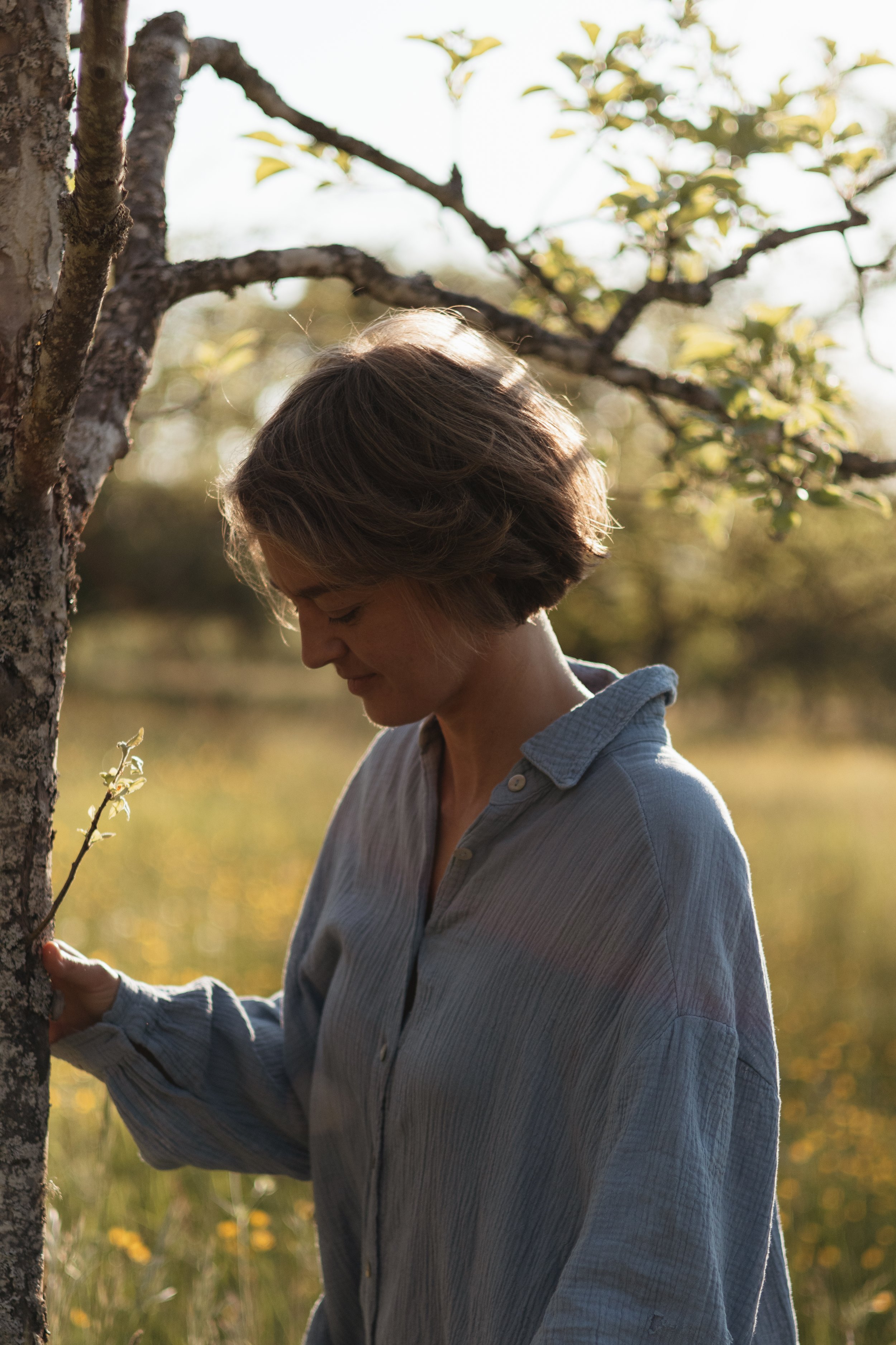 Eine Frau steht im Freien bei Sonnenuntergang neben einem Baum und berührt den Baumstamm, während sie nach unten schaut.