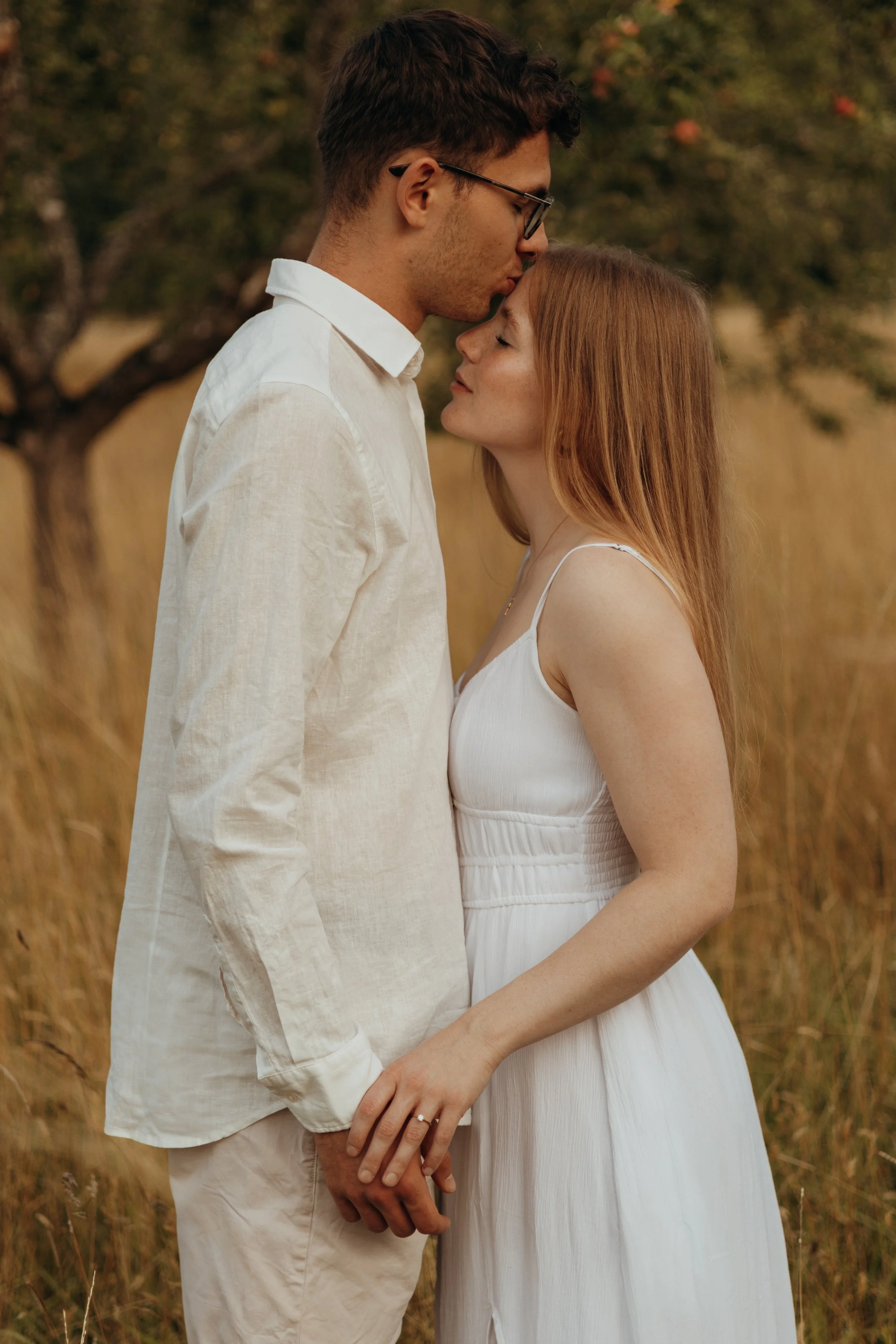 Paar steht in einem Feld, sie halten Händchen, er trägt eine weiße Bluse, sie ein weißes Kleid, sie drücken ihre Stirnen aneinander, es ist wahrscheinlich ein romantisches Fotoshooting.