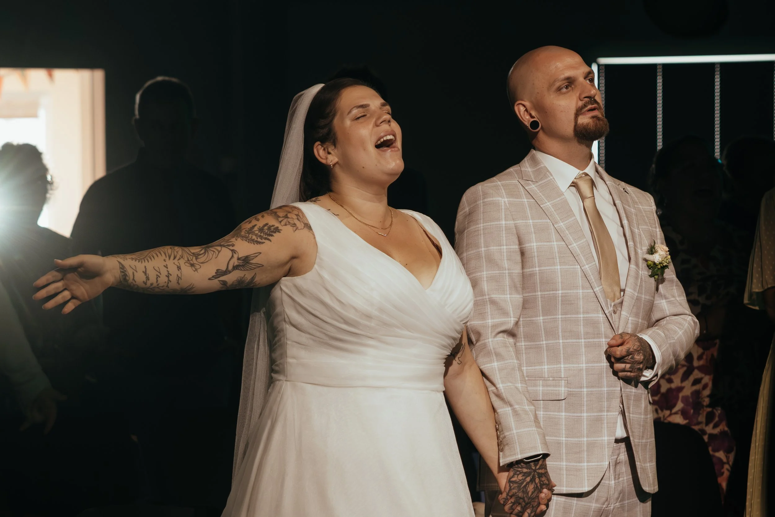 Ein Brautpaar bei einer Hochzeit, die Braut singt oder spricht mit offenem Mund, während der Bräutigam ruhig daneben steht, beide halten sich an den Händen, in einem dunklen Raum mit anderen Gästen im Hintergrund.