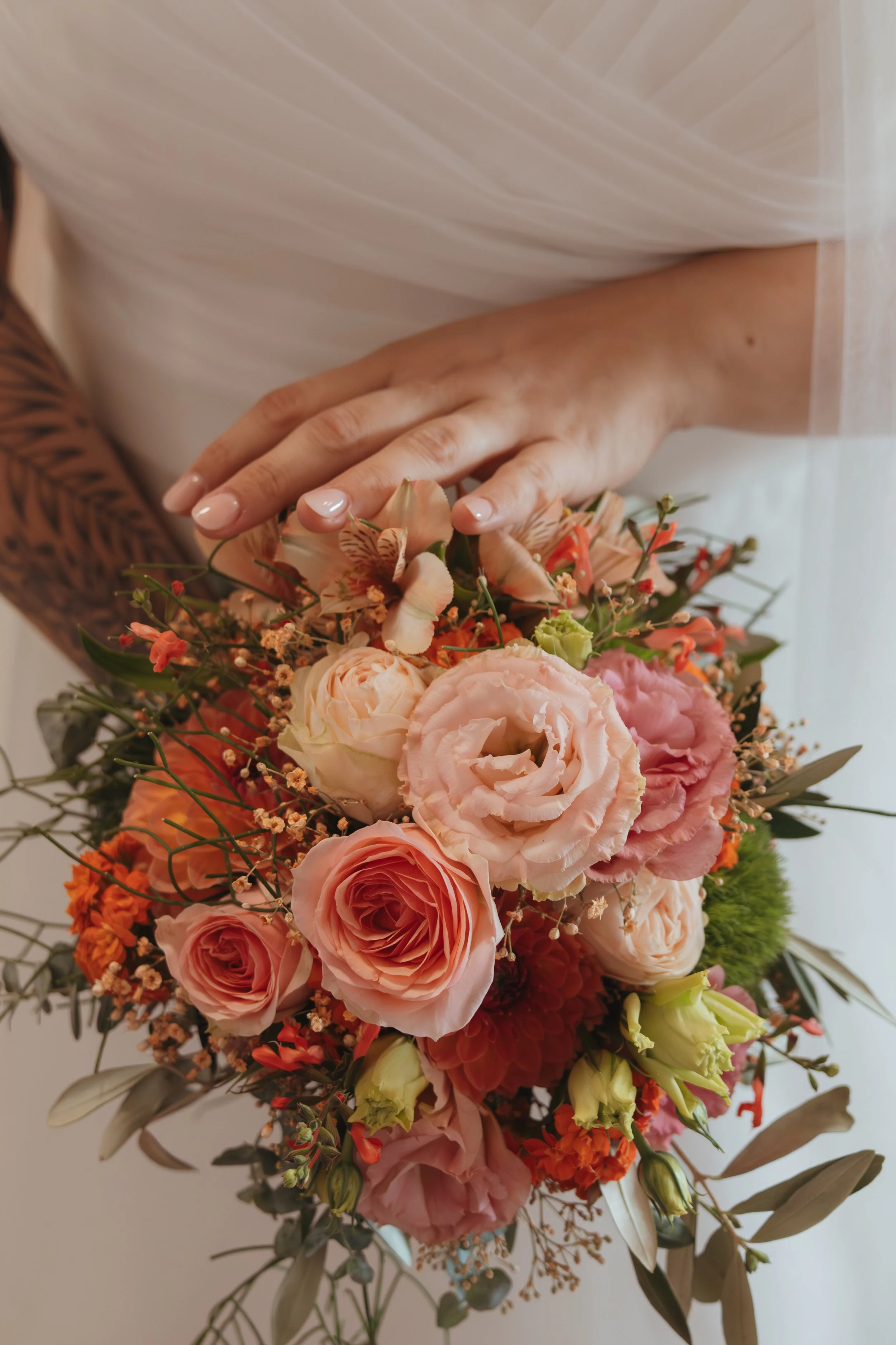 Nahaufnahme eines Brautstraußes mit rosa und peachfarbenen Blumen, gehalten von einer Person mit einer Tätowierung am Arm, vor hellem Hintergrund.