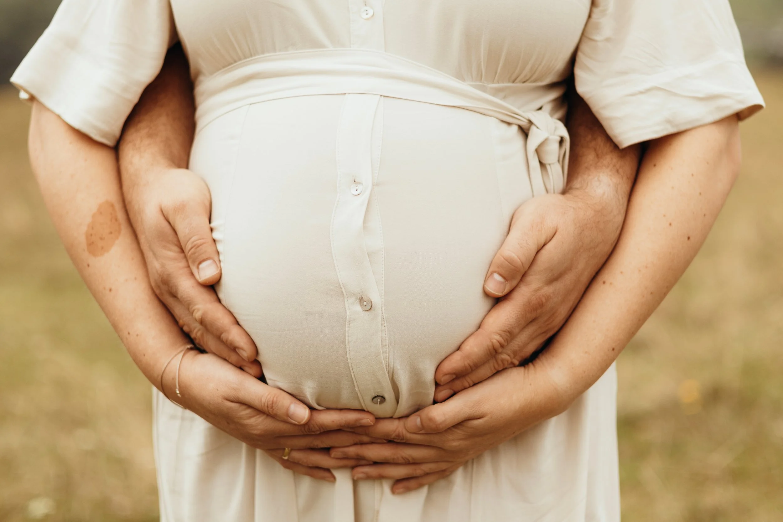 Ein Paar hält die schwangere Bauchseite von einer Frau im beige Kleid, beide haben ihre Hände darauf gelegt.