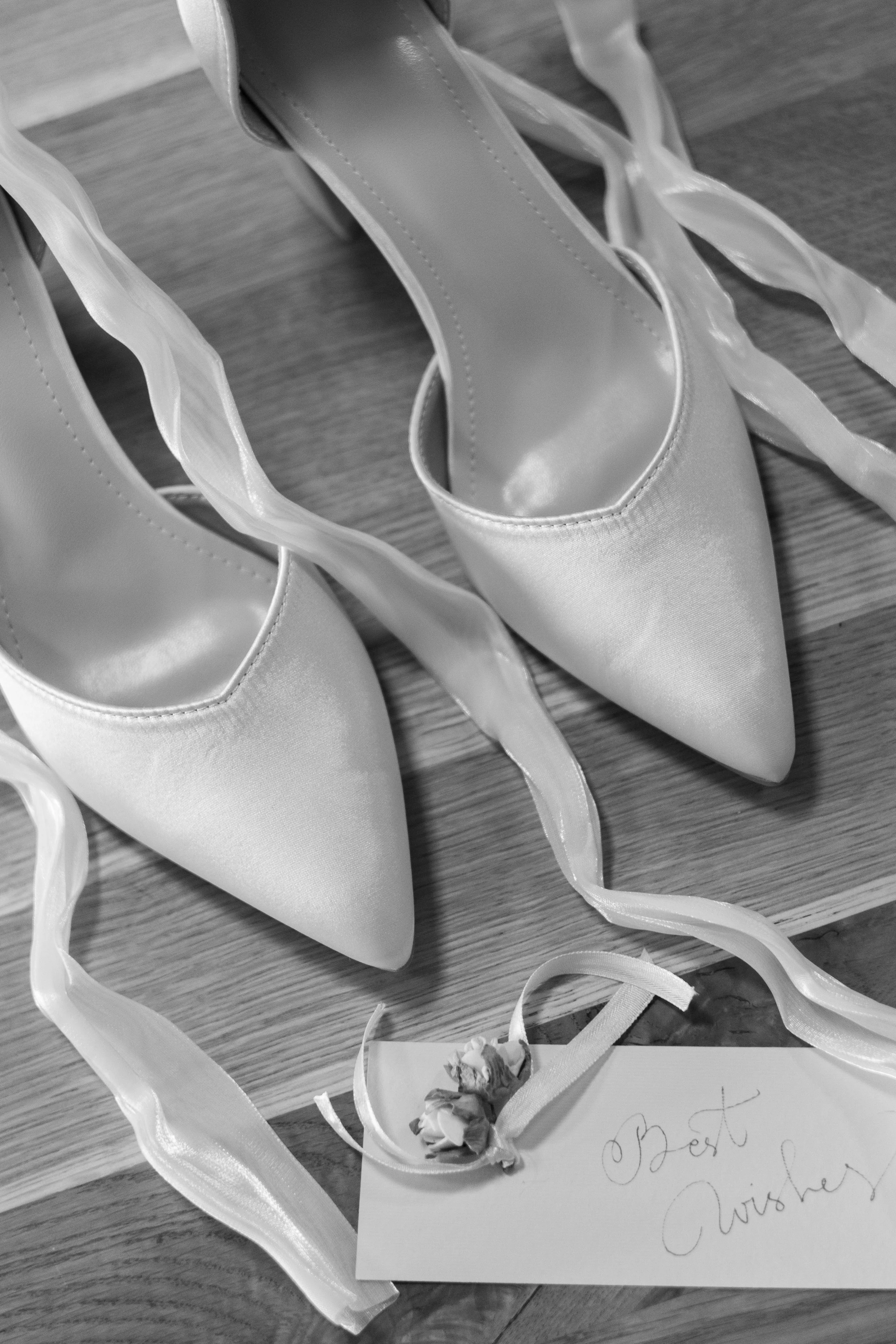 Satin weiße High Heels mit Schleifen und einer handgeschriebenen Karte mit der Aufschrift 'Best Wishes' auf Holztisch.