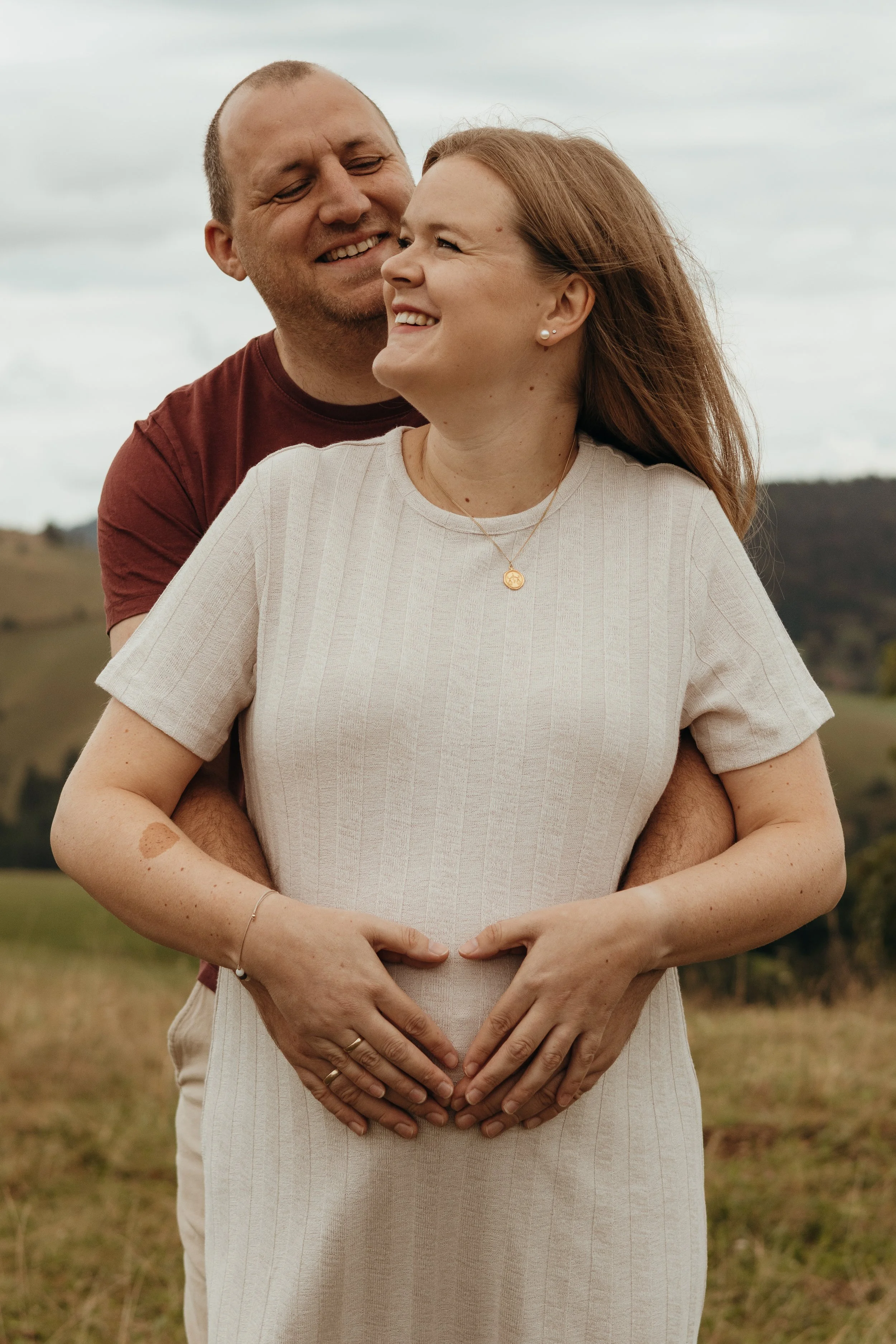 Ein Paar, vermutlich ein schwangerer Frau, steht im Freien in einer ländlichen Umgebung, während der Mann sie von hinten umarmt und beide lächeln.