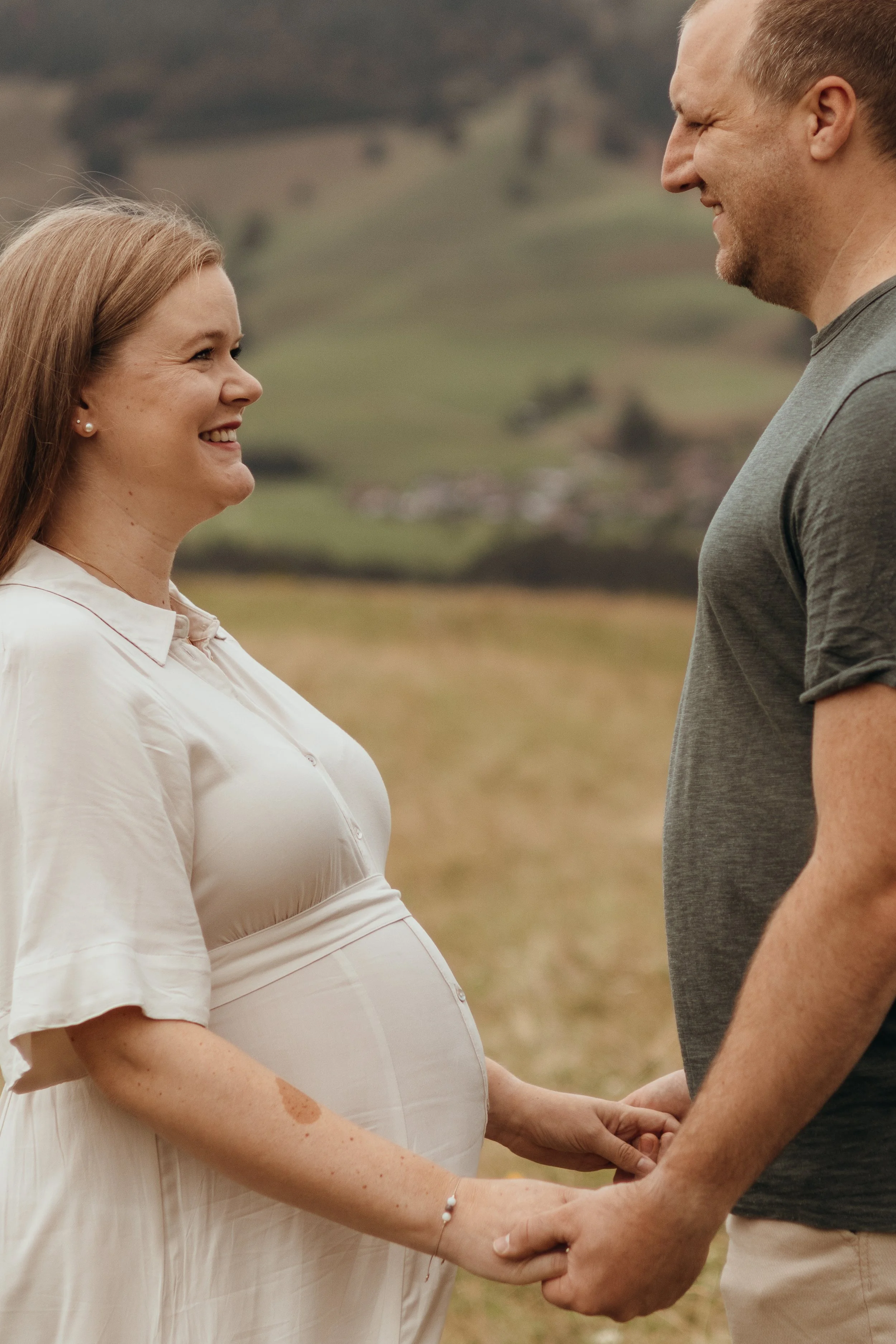 Ein schwangertes Paar hält Händchen und blickt sich verliebt in die Augen, im Freien vor einer ländlichen Landschaft.