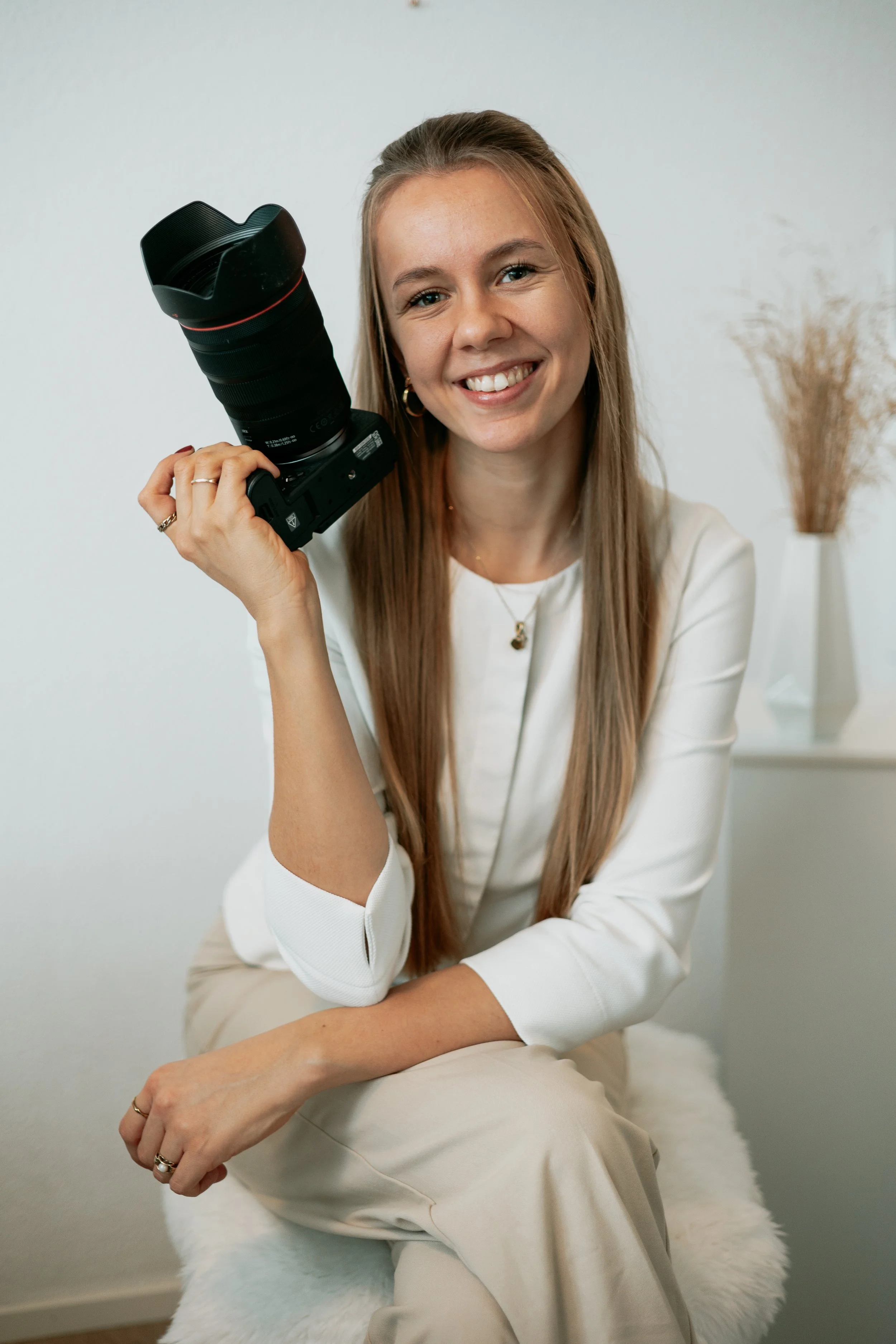 Lächelnde Frau mit langer, glatter brauner Haar hält eine professionelle Kamera mit großem Objektiv und Stativ in der Hand, sitzt auf einem weißen, pelzigen Hocker, im Hintergrund ein kleines Regal mit trockenen Blumen in einer weißen Vase.