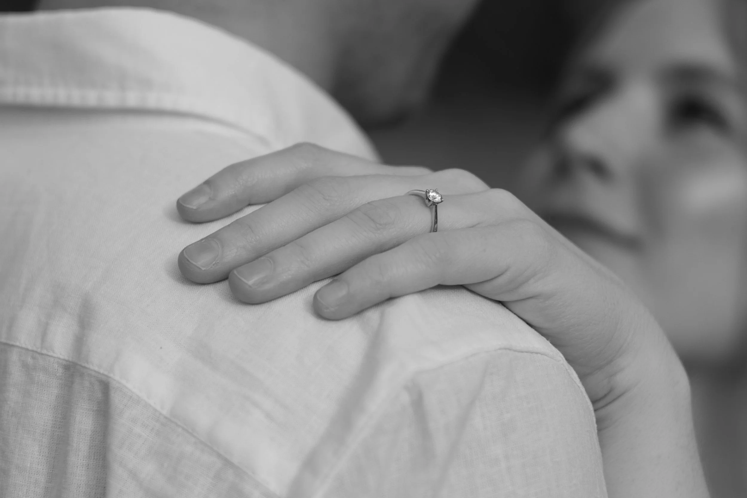 Nahaufnahme einer Hand mit Verlobungsring auf der Schulter eines anderen Menschen, im Hintergrund ein unscharfer Blick auf eine Frau.