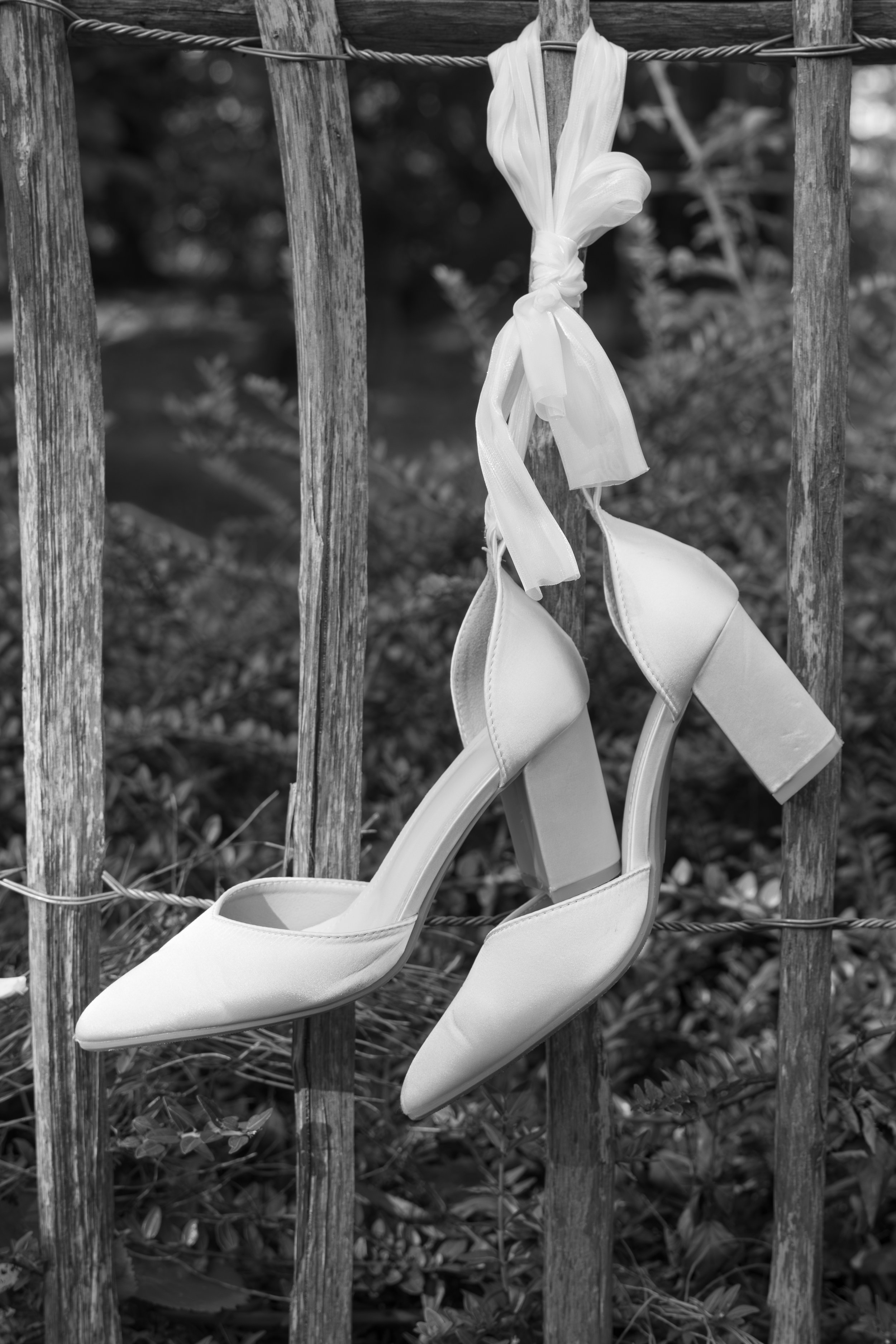 Schwarze und weiße Schuhe mit hohen Absätzen hängen an einem Holzzaun, befestigt mit Schnüren, im Hintergrund sind Blätter und Zweige sichtbar.