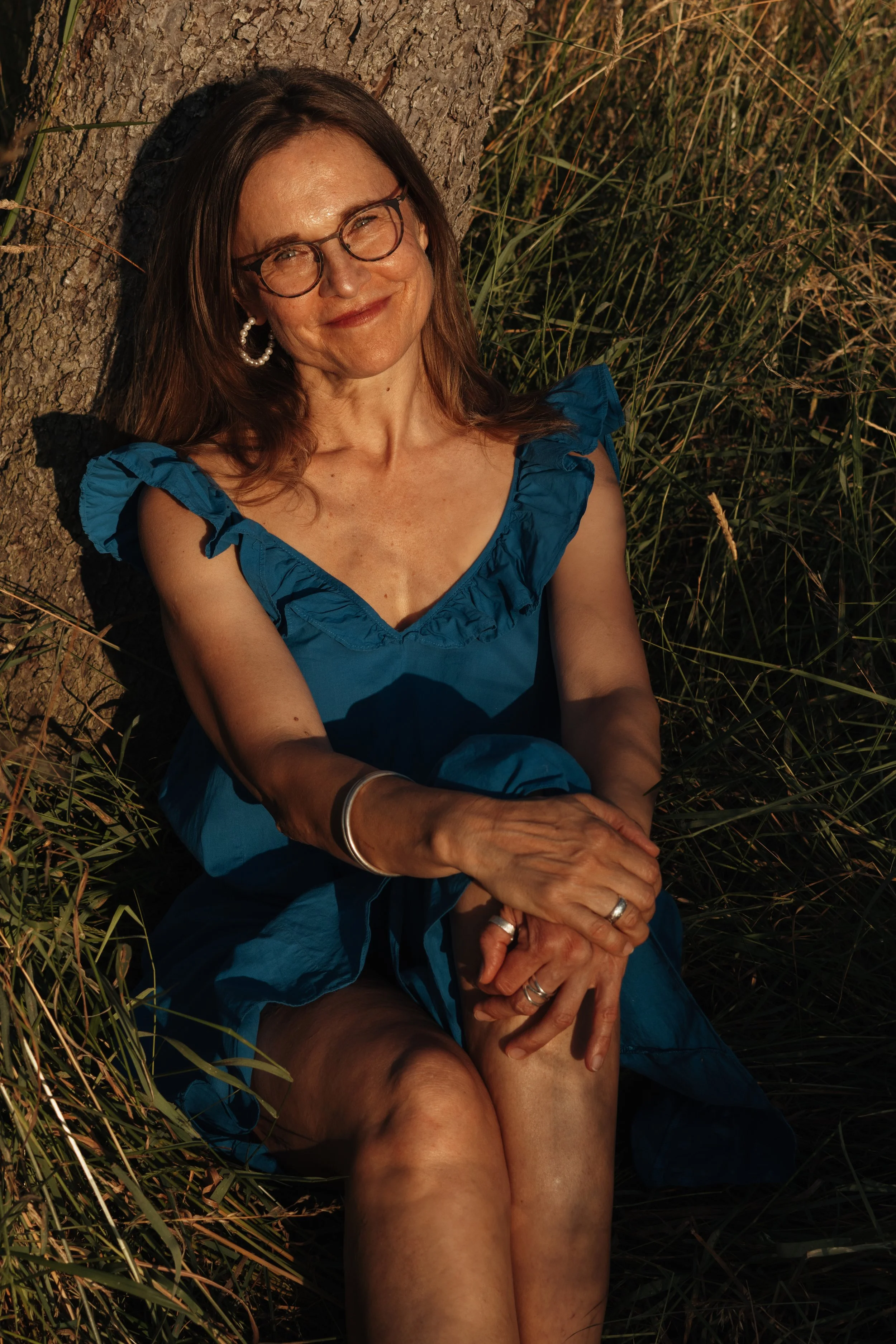 Eine lachende Frau mit Brille und Ohrringen sitzt an einem Baum, umgeben von Gras, bei Sonnenuntergang.