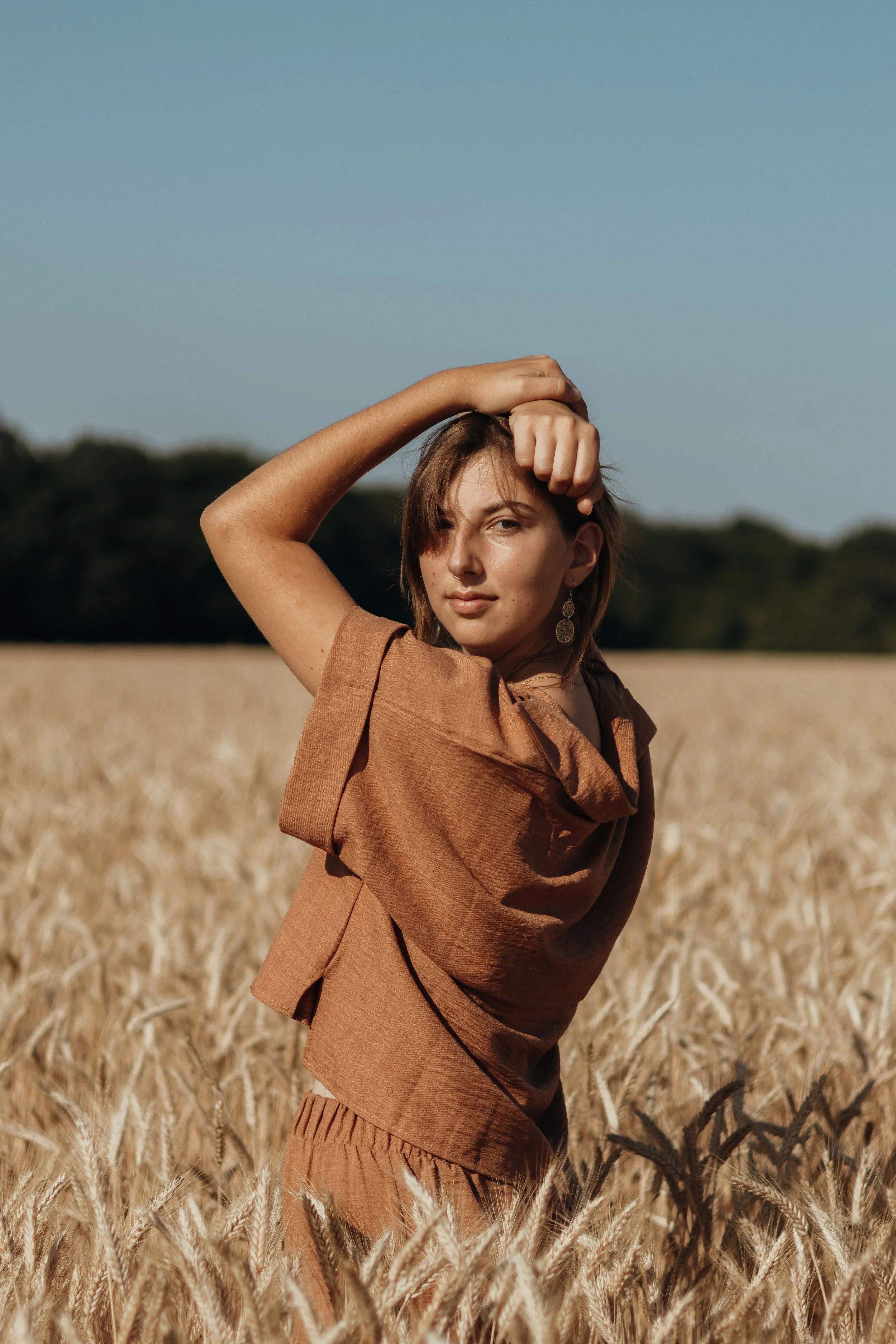 Eine junge Frau steht in einem Weizenfeld bei Sonnenuntergang, trägt braune Kleidung und schaut in die Kamera.