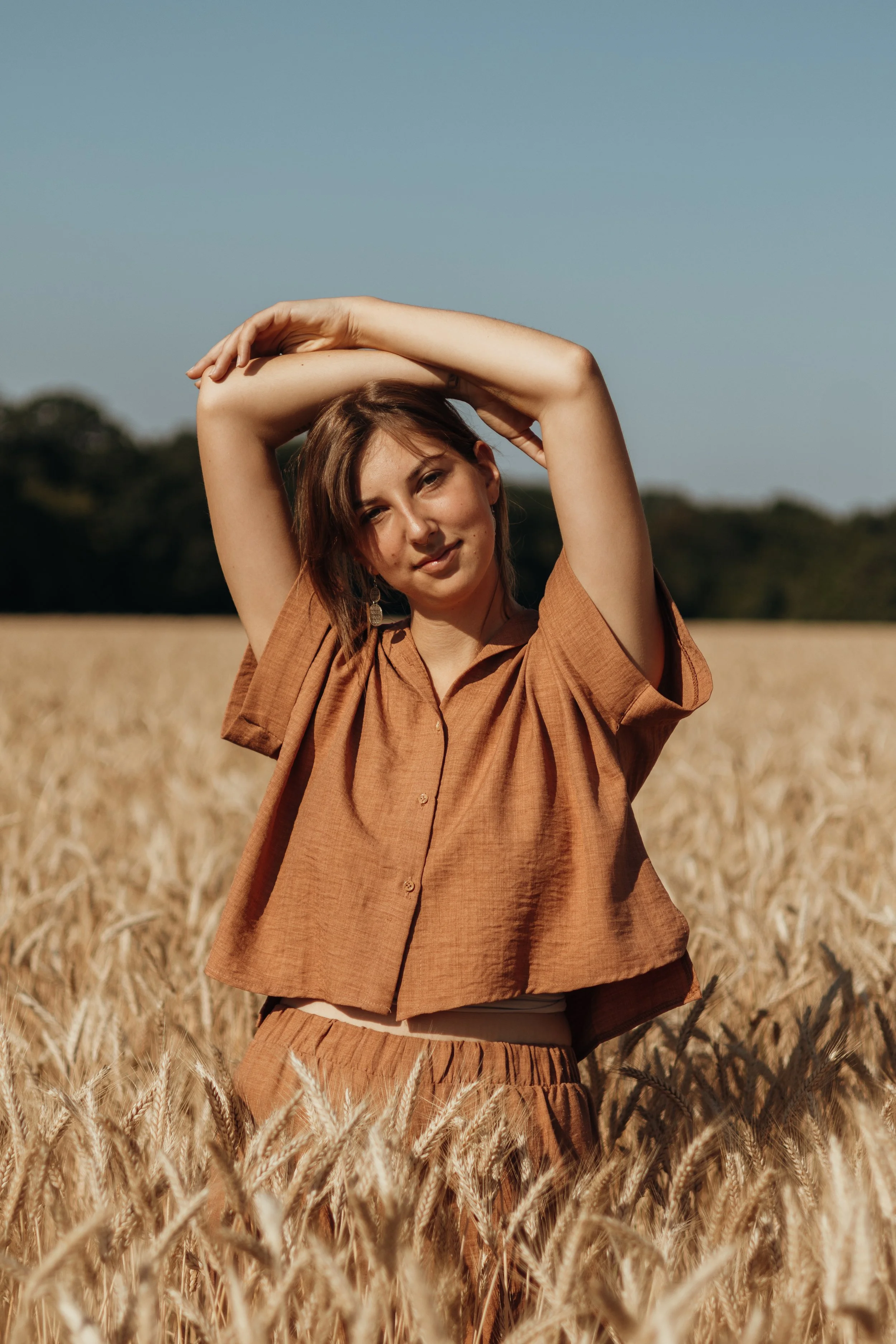 Junge Frau in brauner Hemd und Hose in Weizenfeld bei Sonnenschein, Hände über dem Kopf.