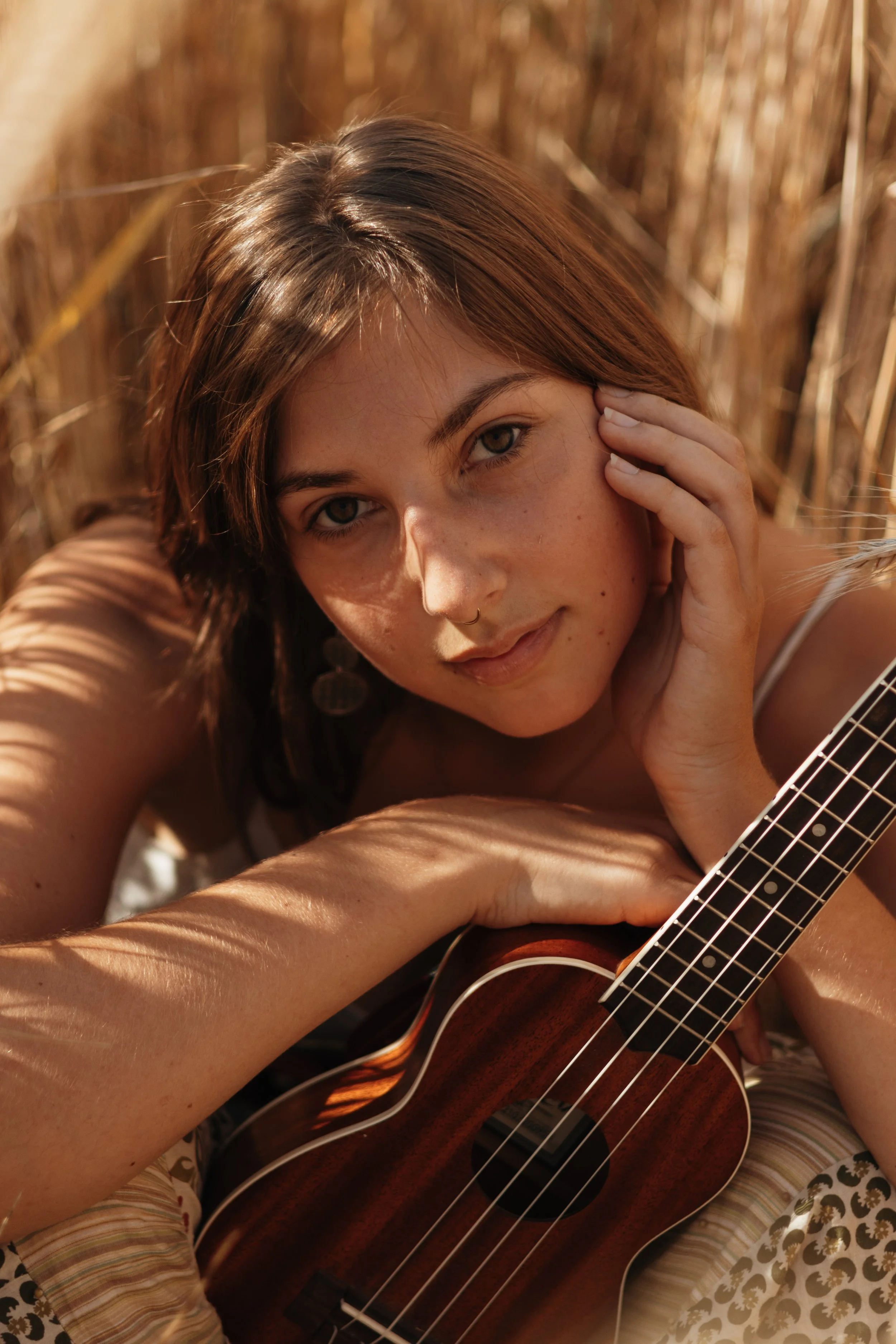 Eine junge Frau mit braunen Haaren und Sommersprossen liegt in einem Feld aus Schilf mit einer Gitarre, schaut in die Kamera und berührt ihr Gesicht mit ihrer Hand.
