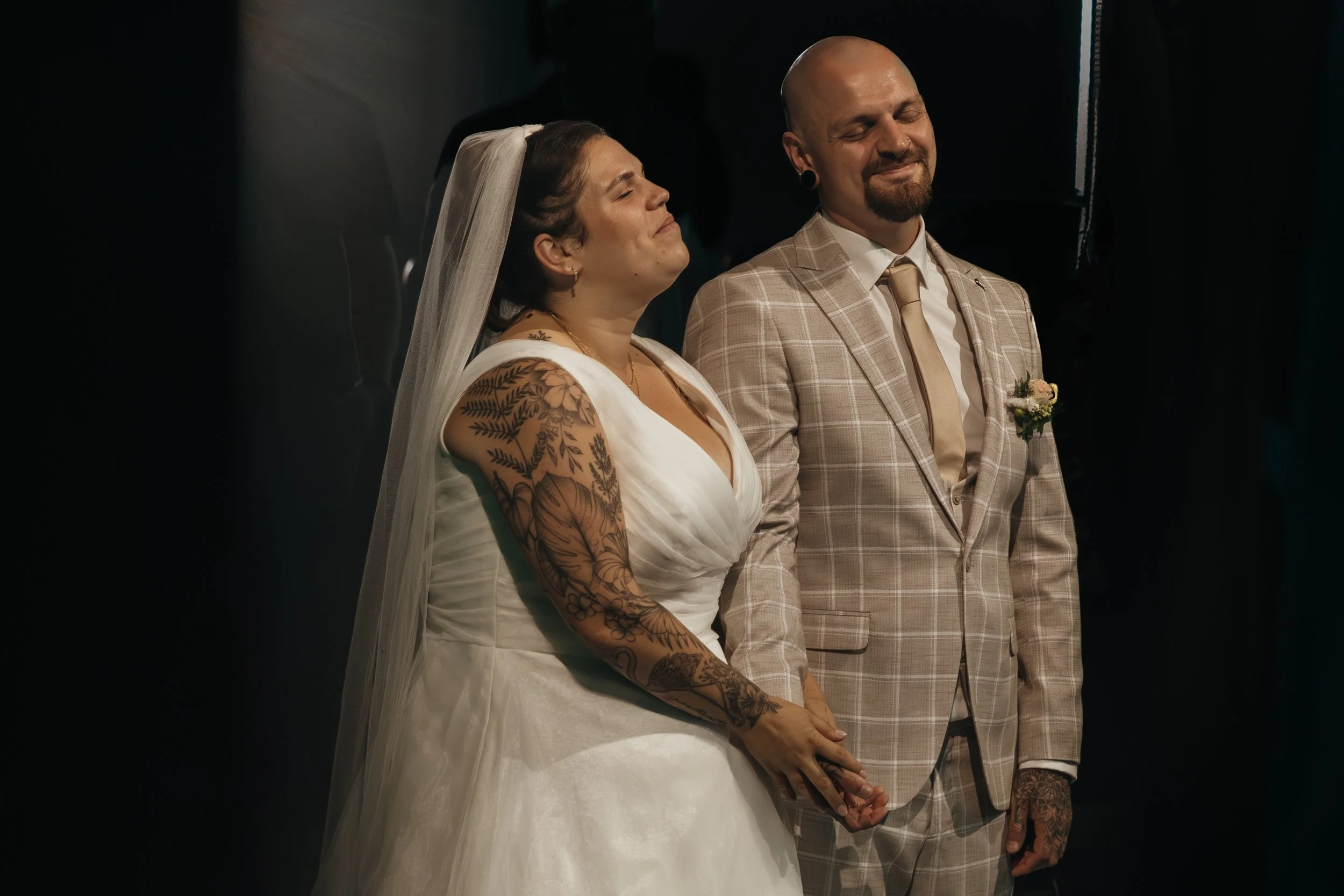 Ein Brautpaar beim Ja-Wort, die Braut trägt ein weißes Hochzeitskleid mit Tattoos am Arm, während der Bräutigam einen beige karierten Anzug trägt, beide halten sich an den Händen, in einem dunklen Raum.