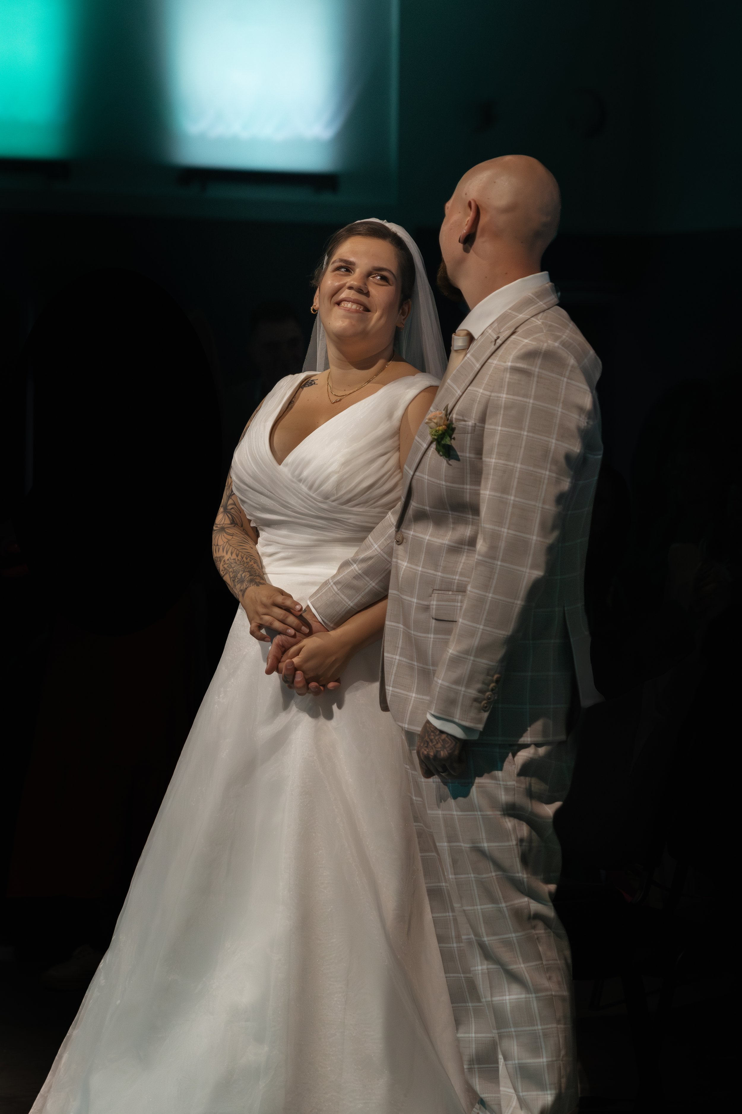 Ein Brautpaar steht während der Hochzeit, die Braut trägt ein weißes Kleid und einen Schleier, der Bräutigam trägt einen beige karierten Anzug. Sie halten sich an den Händen, die Braut lächelt glücklich.