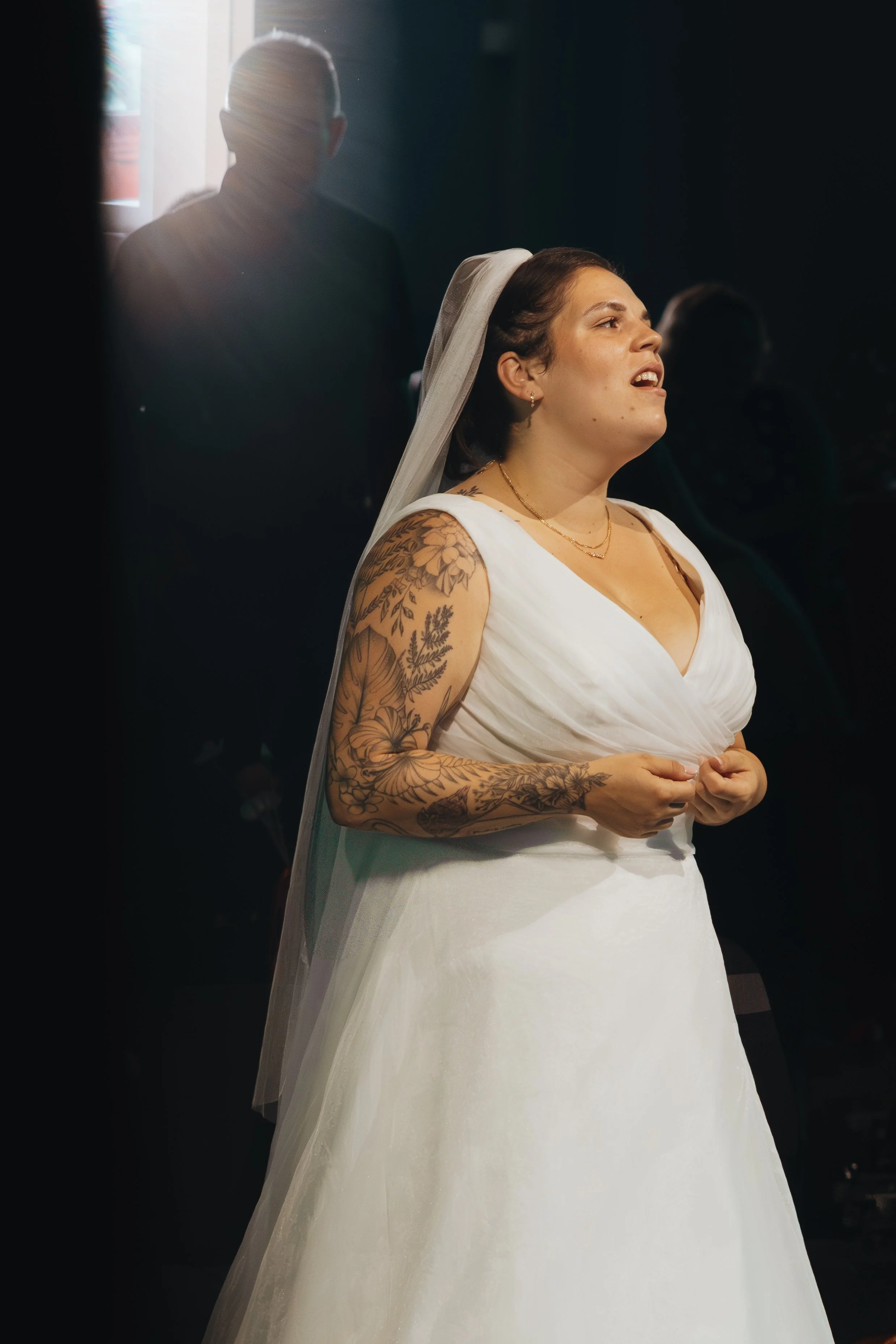 Eine Braut in einem weißen Kleid mit tätowierten Armen, die an einer Hochzeit beteiligt ist. Im Hintergrund ist eine Person in dunkler Kleidung sichtbar.